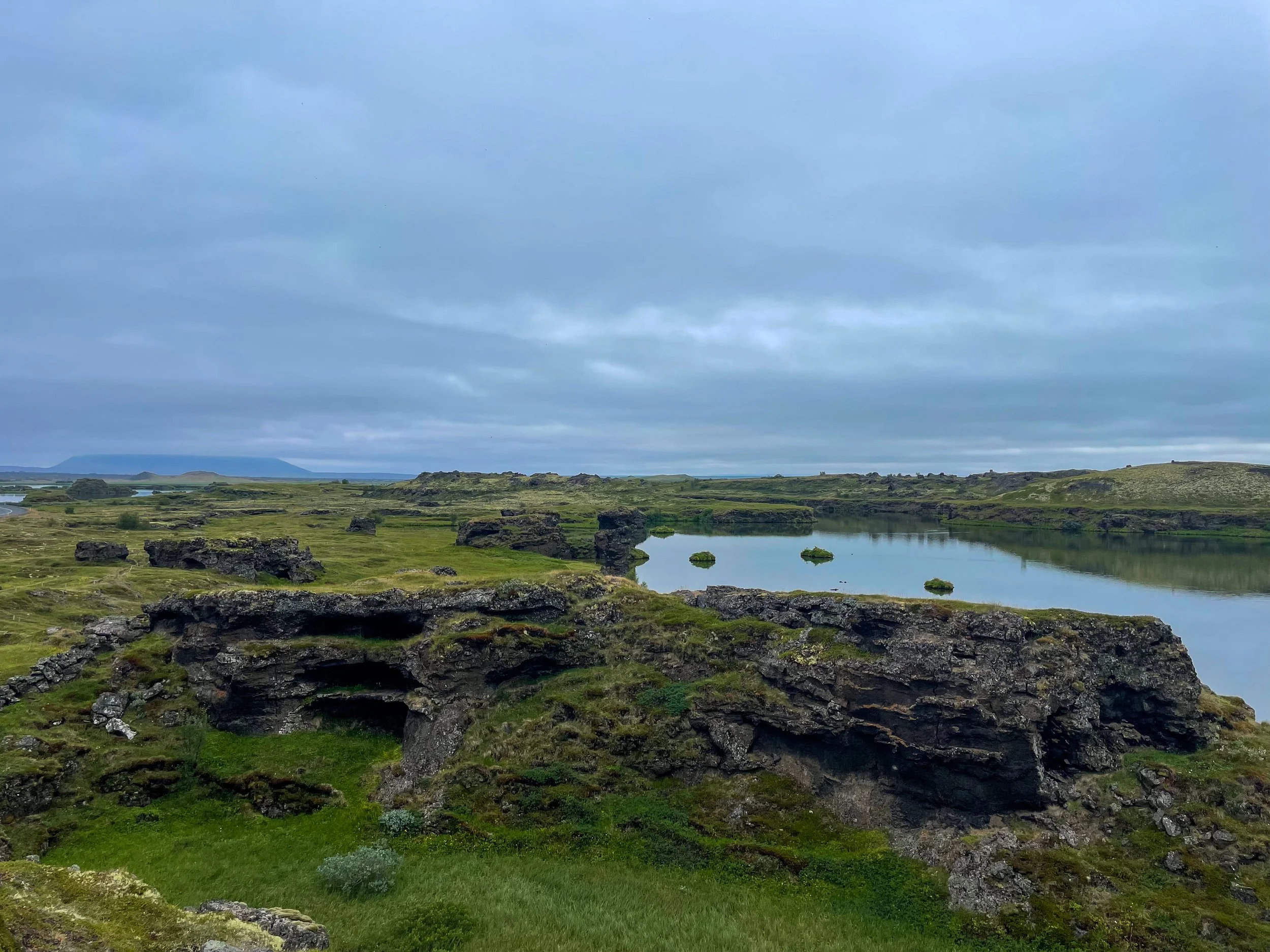 Icelandic landscape with moss-covered rocks, a calm lake, and rolling green hills under a cloudy sky.