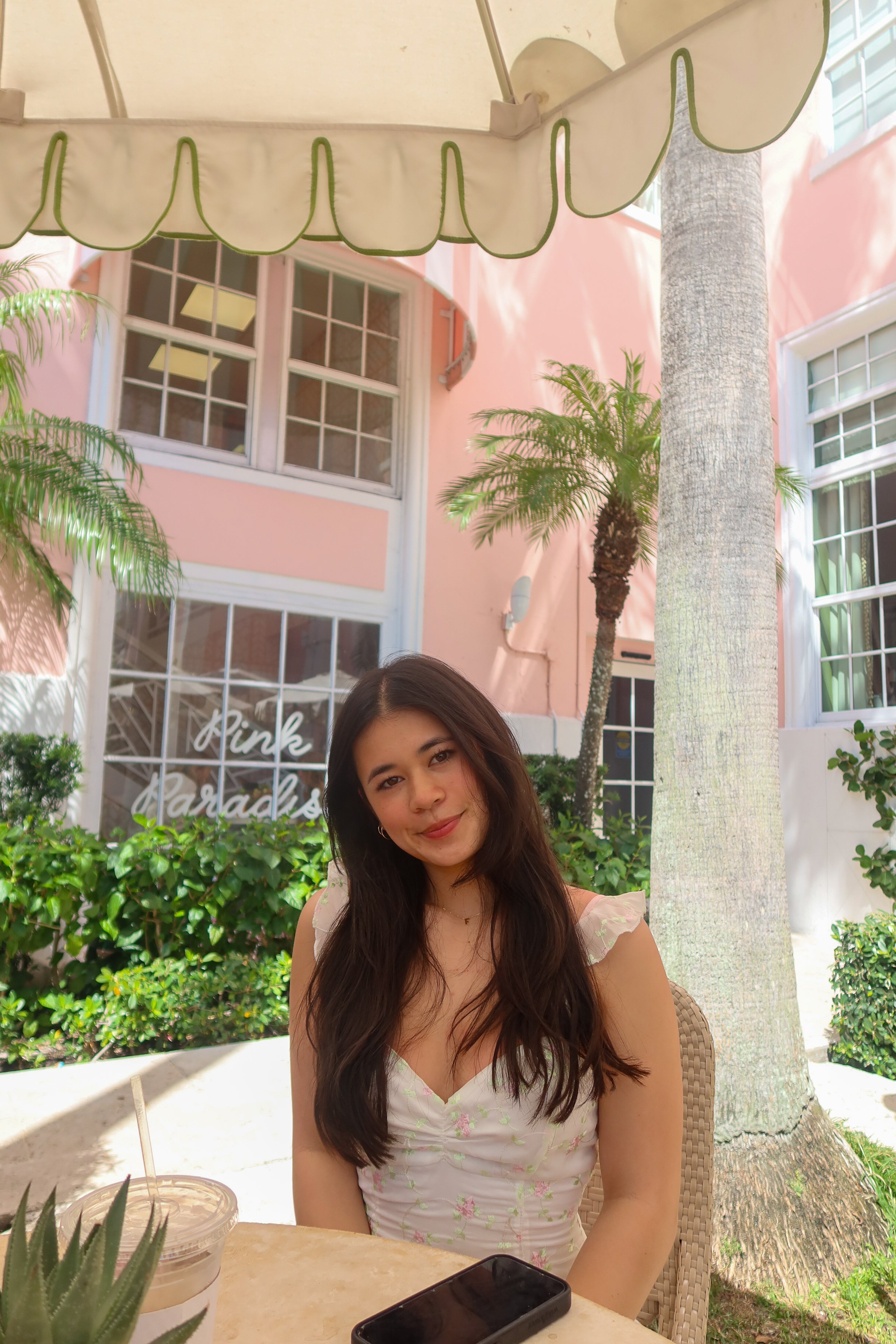 A young woman with long dark hair smiles at the camera while sitting at an outdoor cafe table in a garden area with lush greenery and palm trees, under a white canopy.