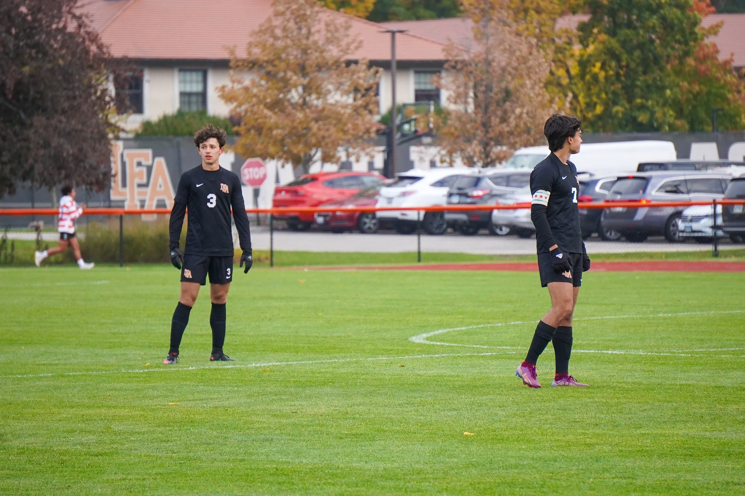 Two soccer players wearing black uniforms stand on a green field, with trees, parked cars, and a building with a red roof in the background.