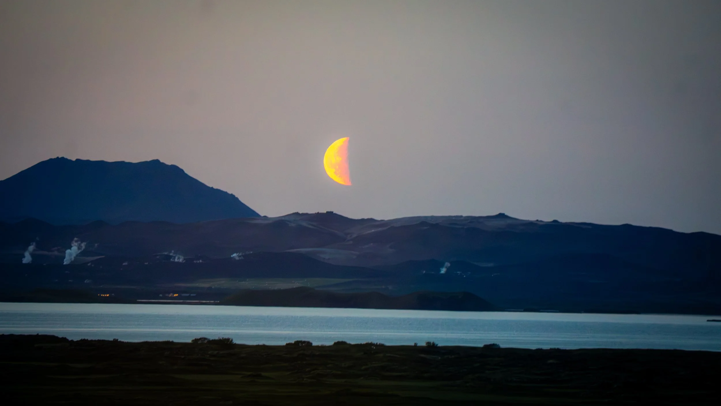 A partially eclipsed moon rises behind mountains at dusk, with a body of water in the foreground and smoke rising from chimneys.