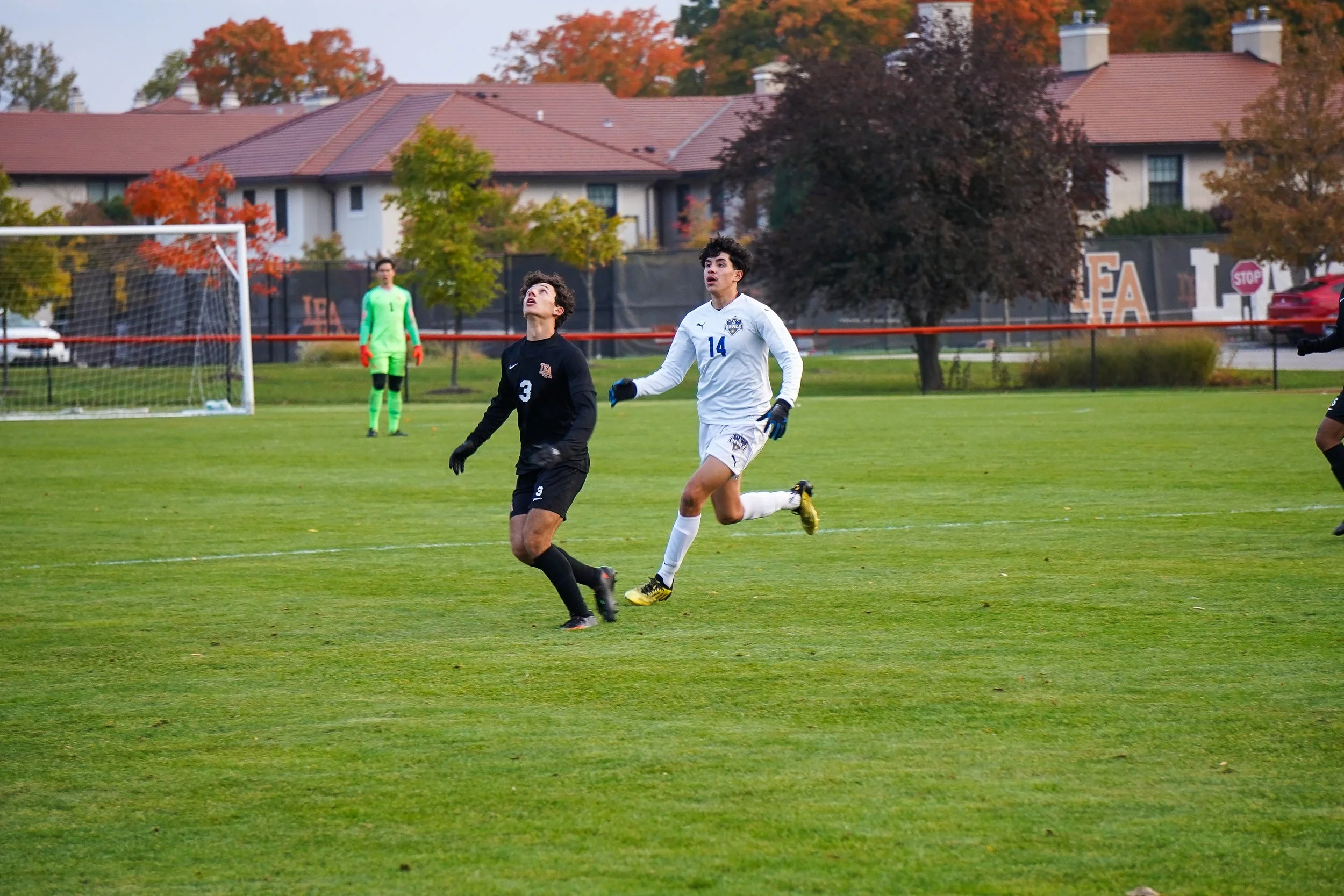 Two soccer players on a field, one in black and one in white, chasing the ball, with a goalkeeper in green and red in the background.