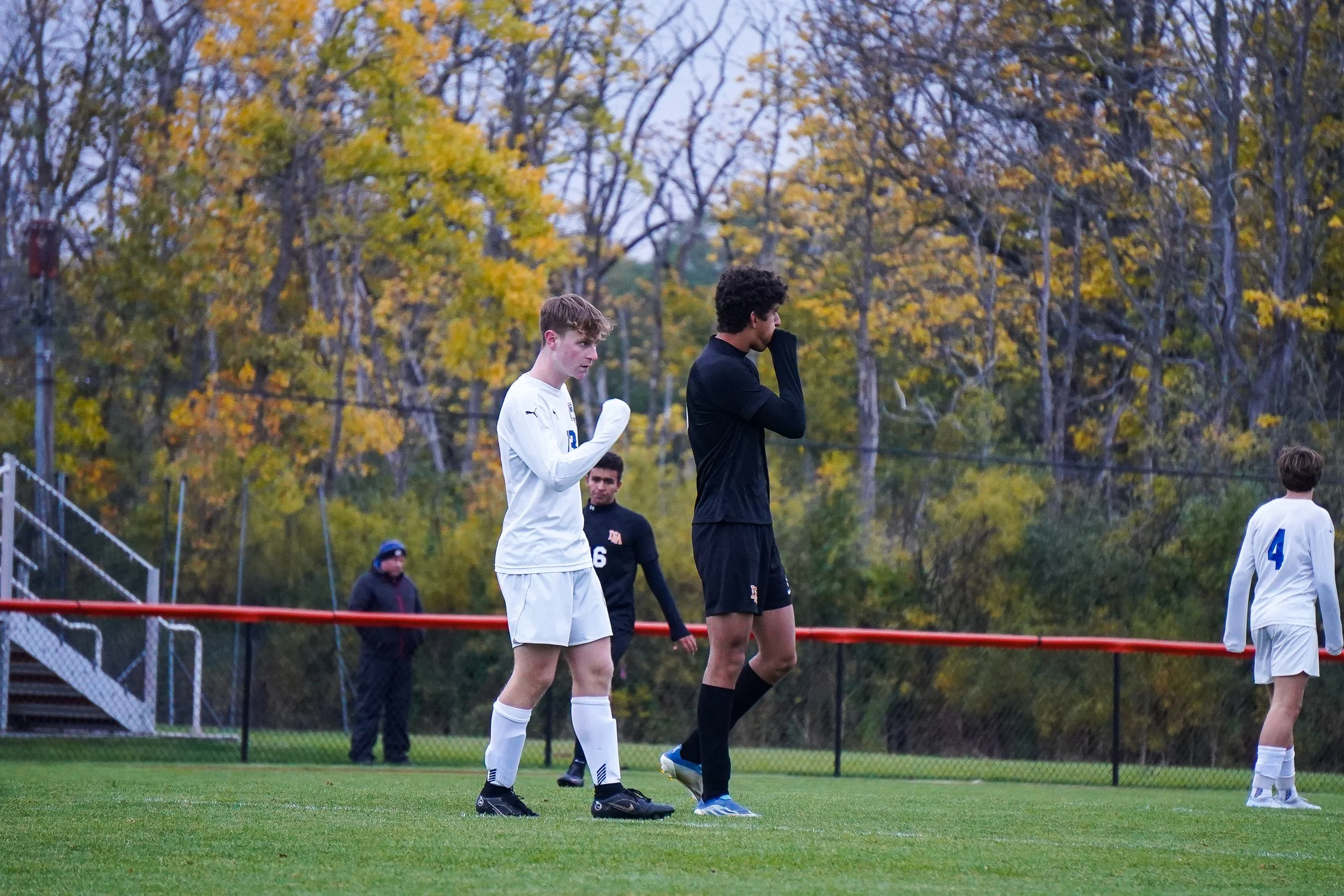 Two soccer players stretching on a field, with a background of autumn trees and a person walking along the sideline.