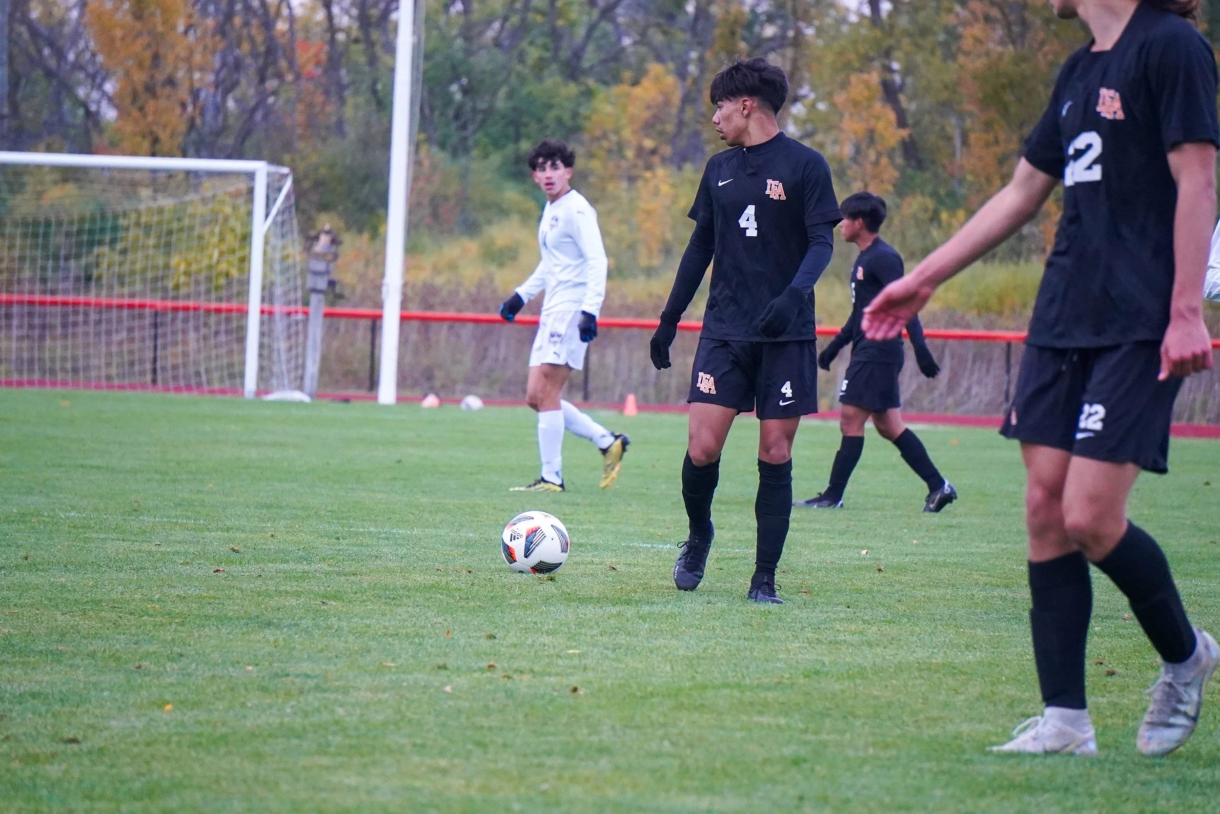 Soccer players on the field preparing for play, with some players wearing black uniforms and one in white, on a green grass field in a park-like setting with trees in autumn colors in the background.