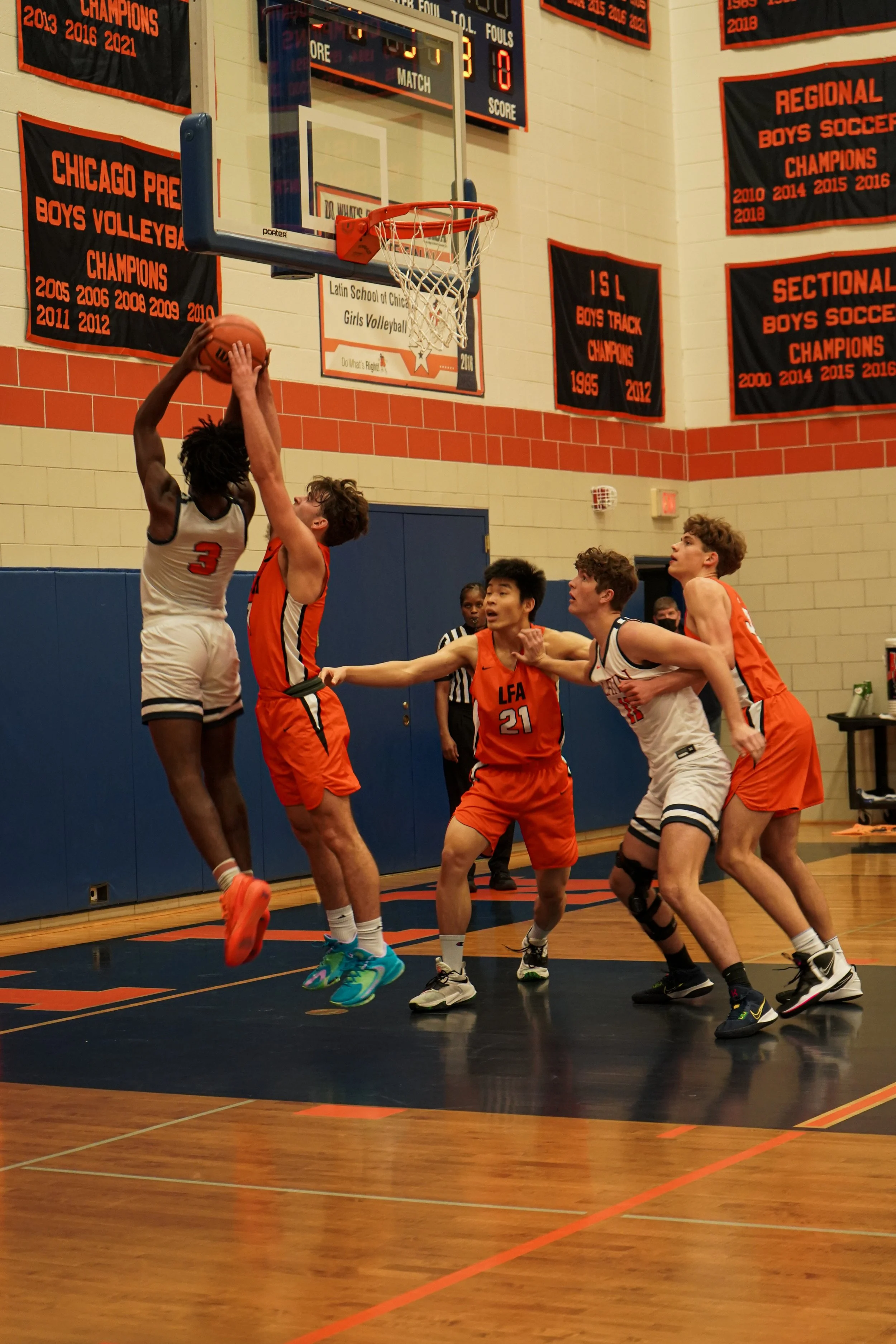 Young basketball players compete near the basket during a game in a gymnasium. One player is jumping to make a shot while others are trying to block or defend. Banners with team achievements hang on the wall behind.