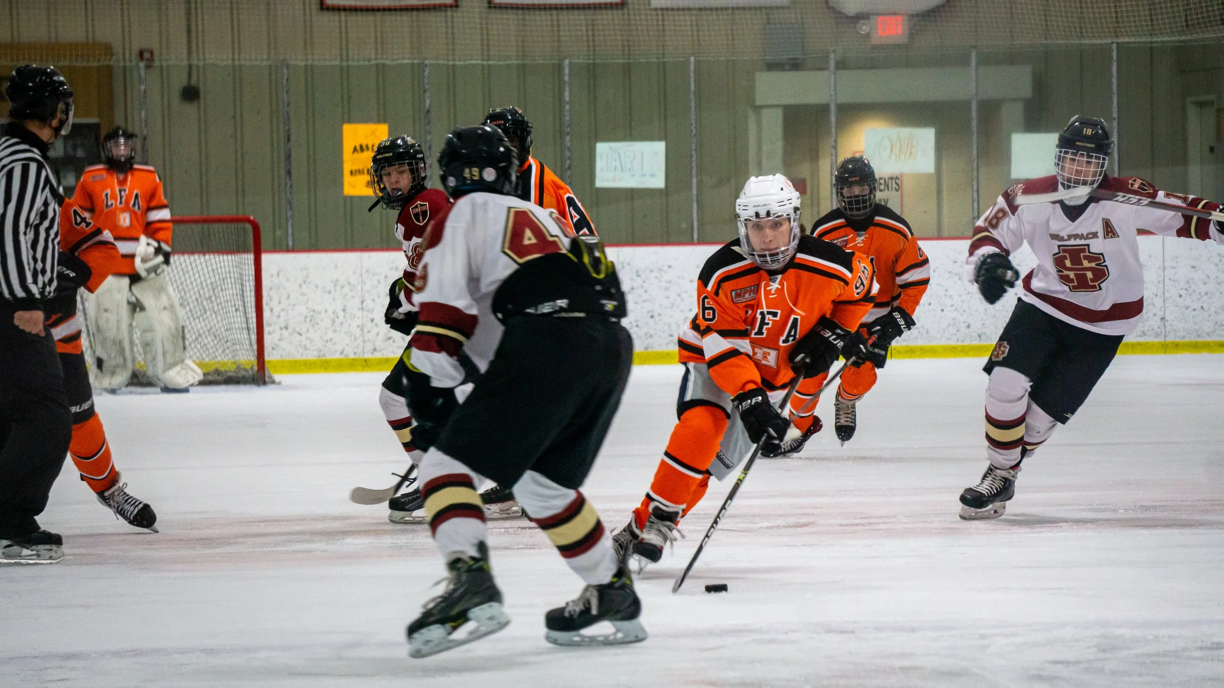 Ice hockey game with players in orange and white jerseys, a referee, and a goalie on the ice rink.