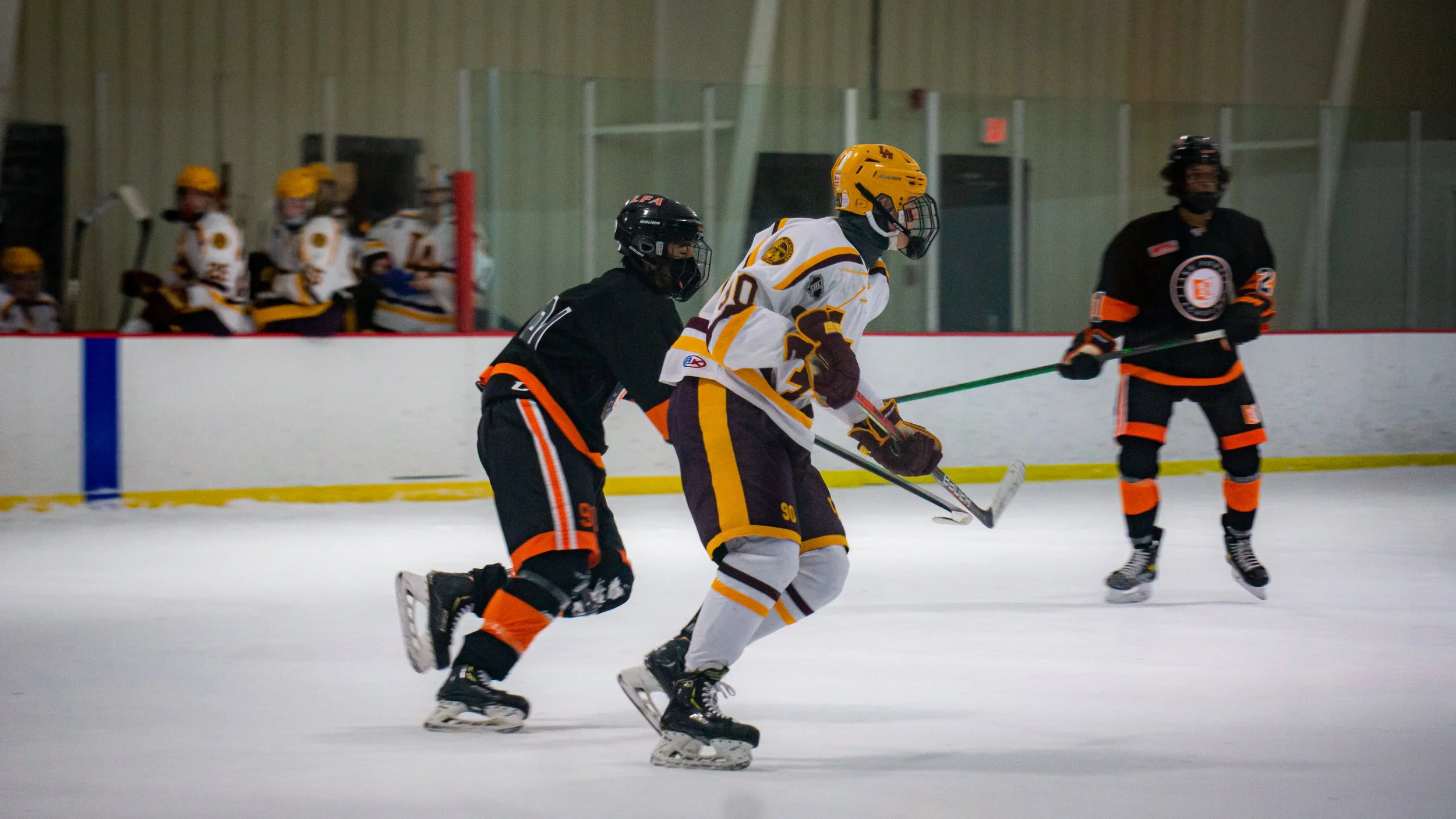 Hockey players on the ice during a game, with players in black and orange jerseys and one in a white and purple jersey, with team benches and players in yellow helmets in the background.