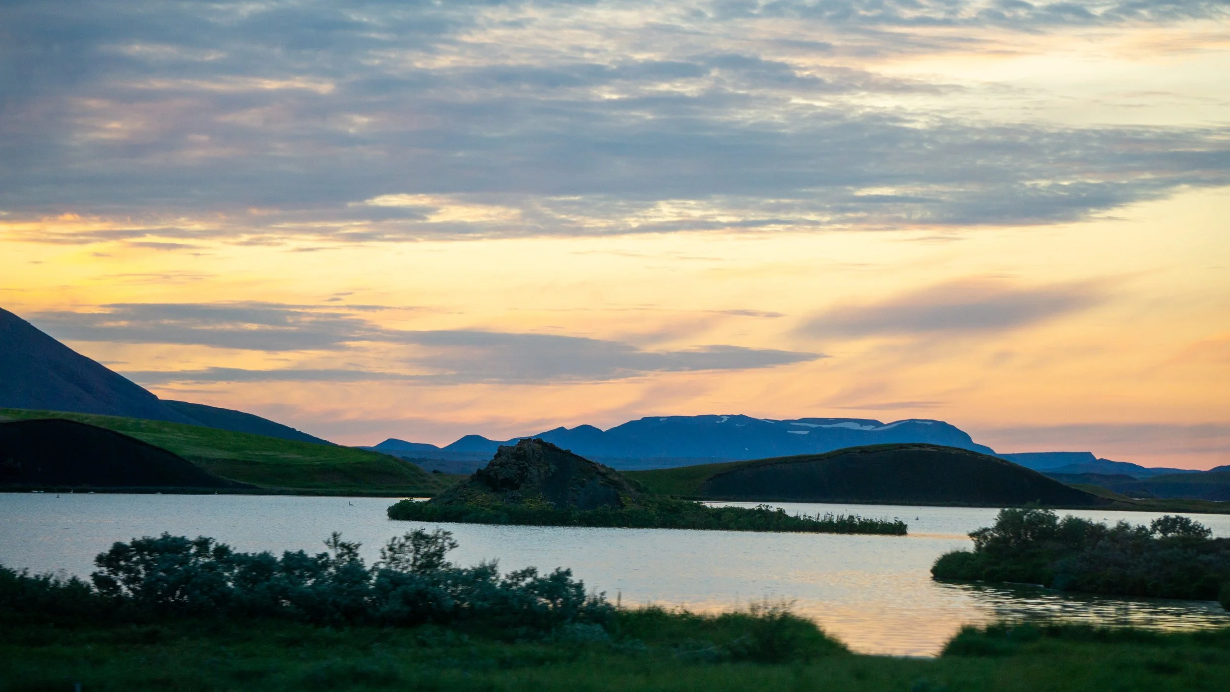 Landscape of a lake with small islands, surrounded by green hills and mountains during sunset with clouds in the sky.