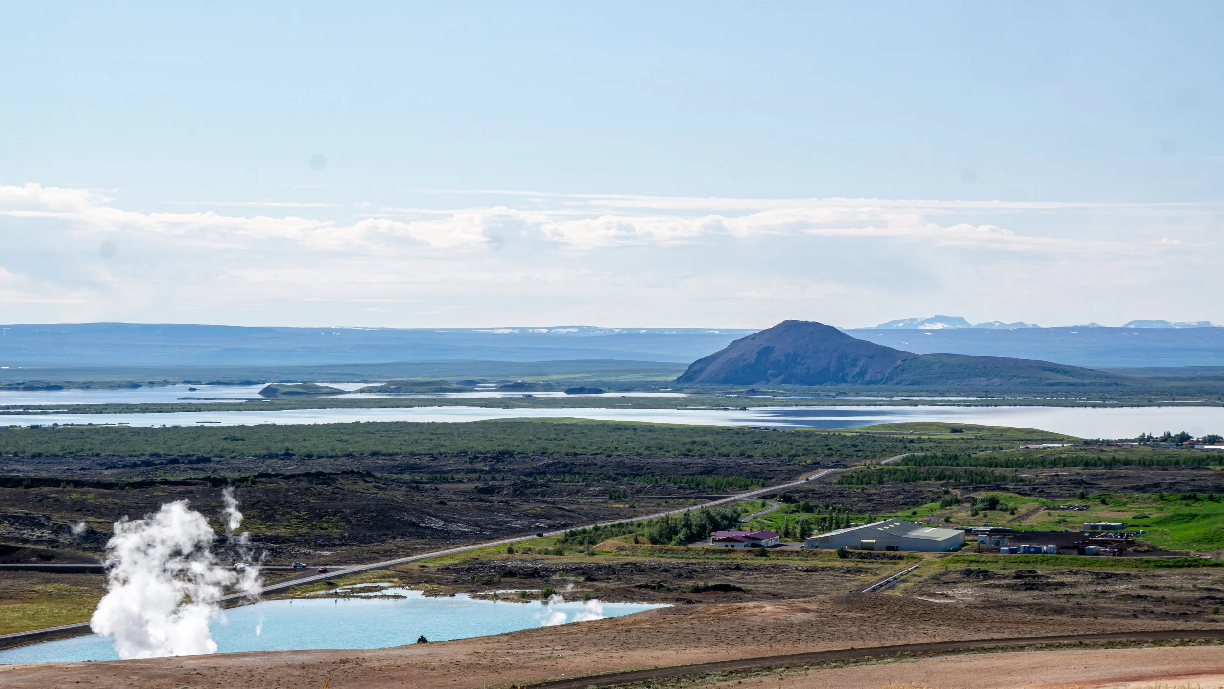 Scenic landscape with geothermal hot spring with steam rising, a winding road, green fields, a large mountain, and a body of water in the background under a partly cloudy sky.