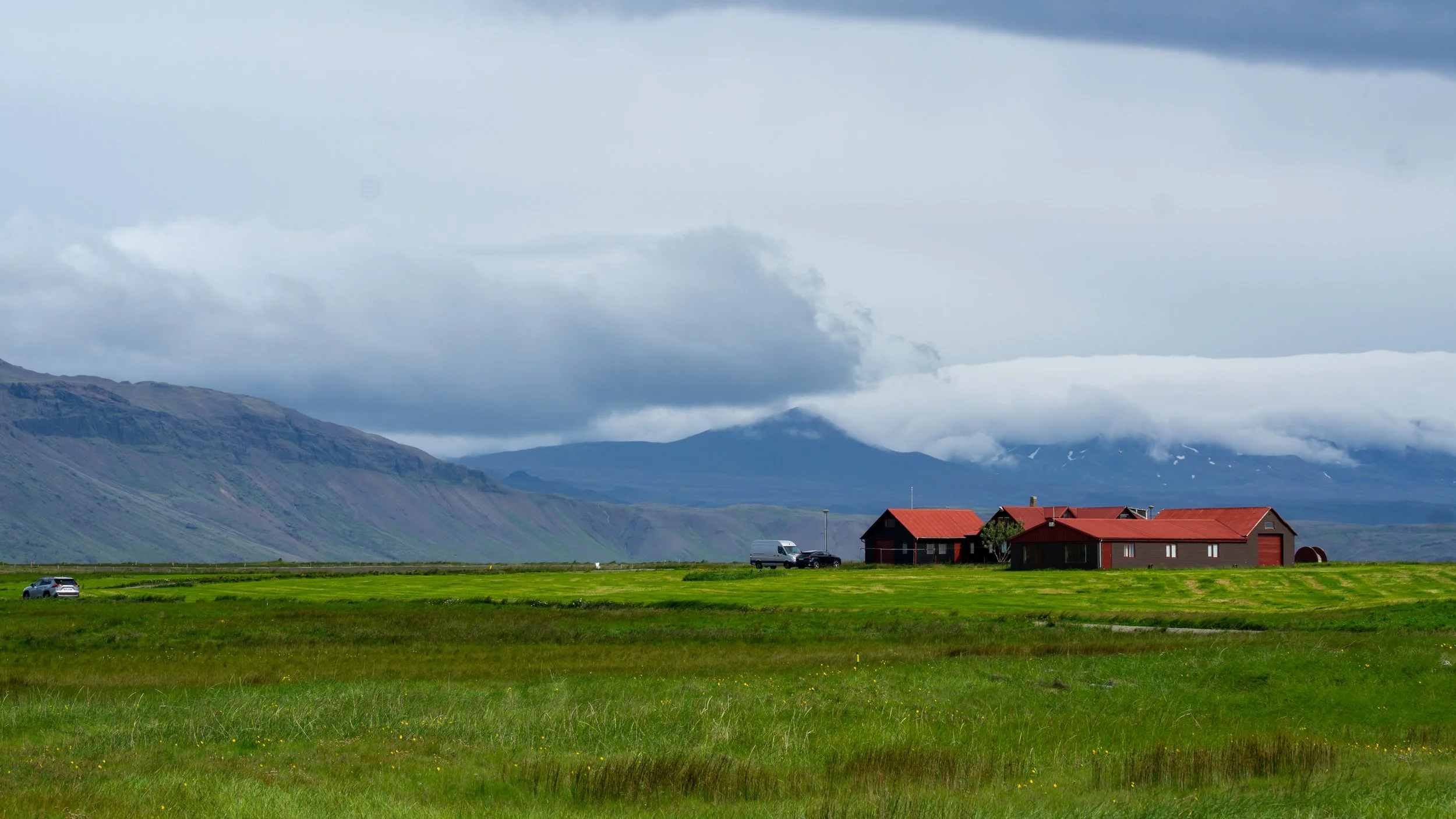 A rural landscape with green fields, a few dark red buildings, mountains in the background, and a cloudy sky.