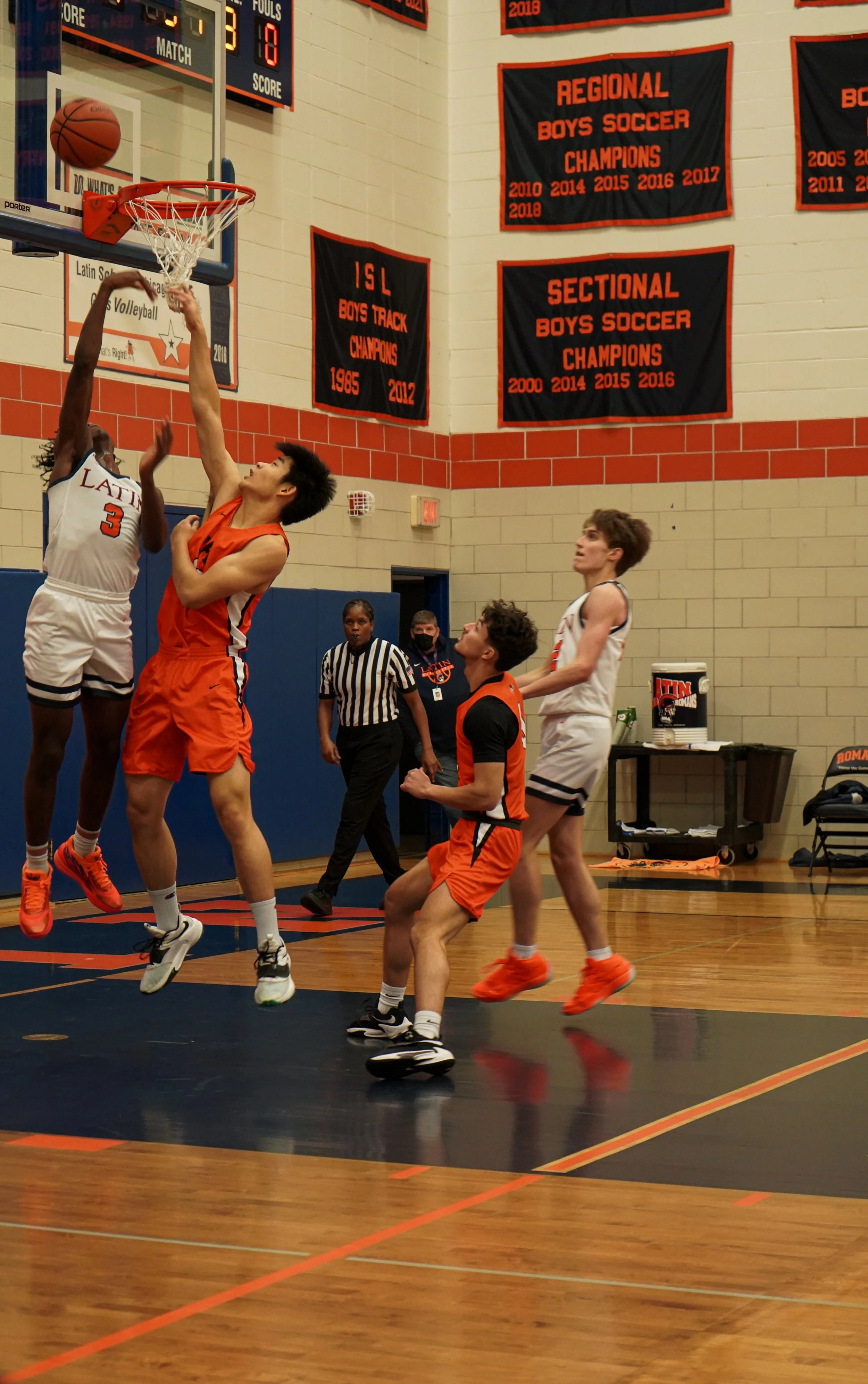 A basketball game in progress with players from two teams, one in orange jerseys and one in white jerseys, jumping and reaching for the ball near the hoop. An official and other players observe on the court.