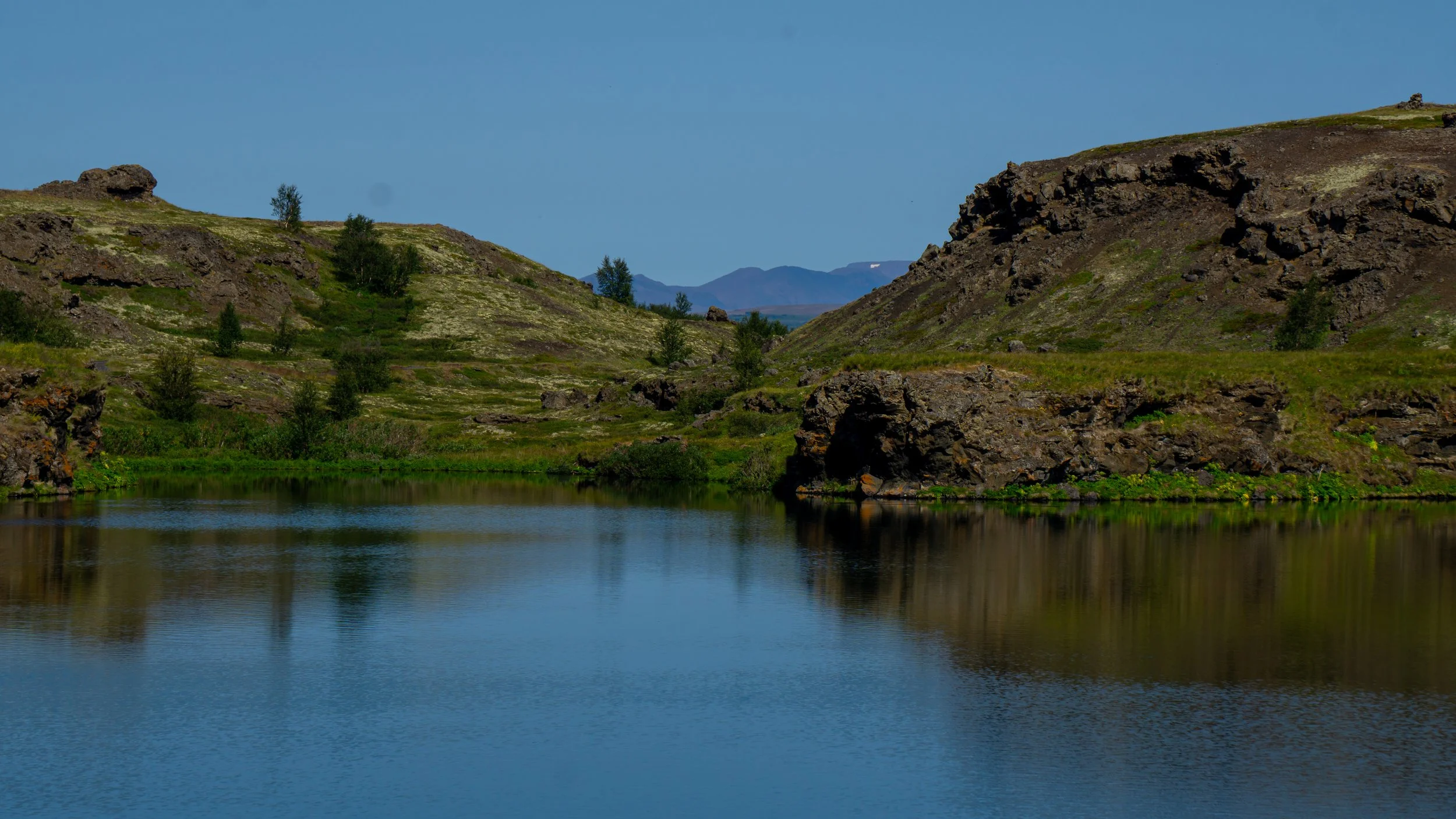 A peaceful river flowing through rocky hills with green vegetation and distant mountains under a clear blue sky.