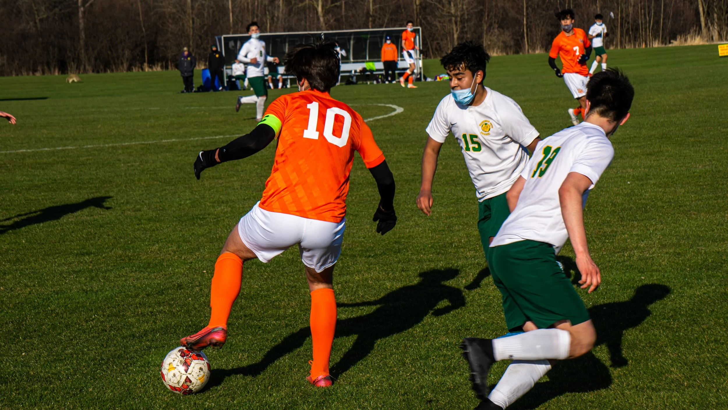 Soccer match with players in orange and white uniforms on a grassy field, with some players wearing face masks.