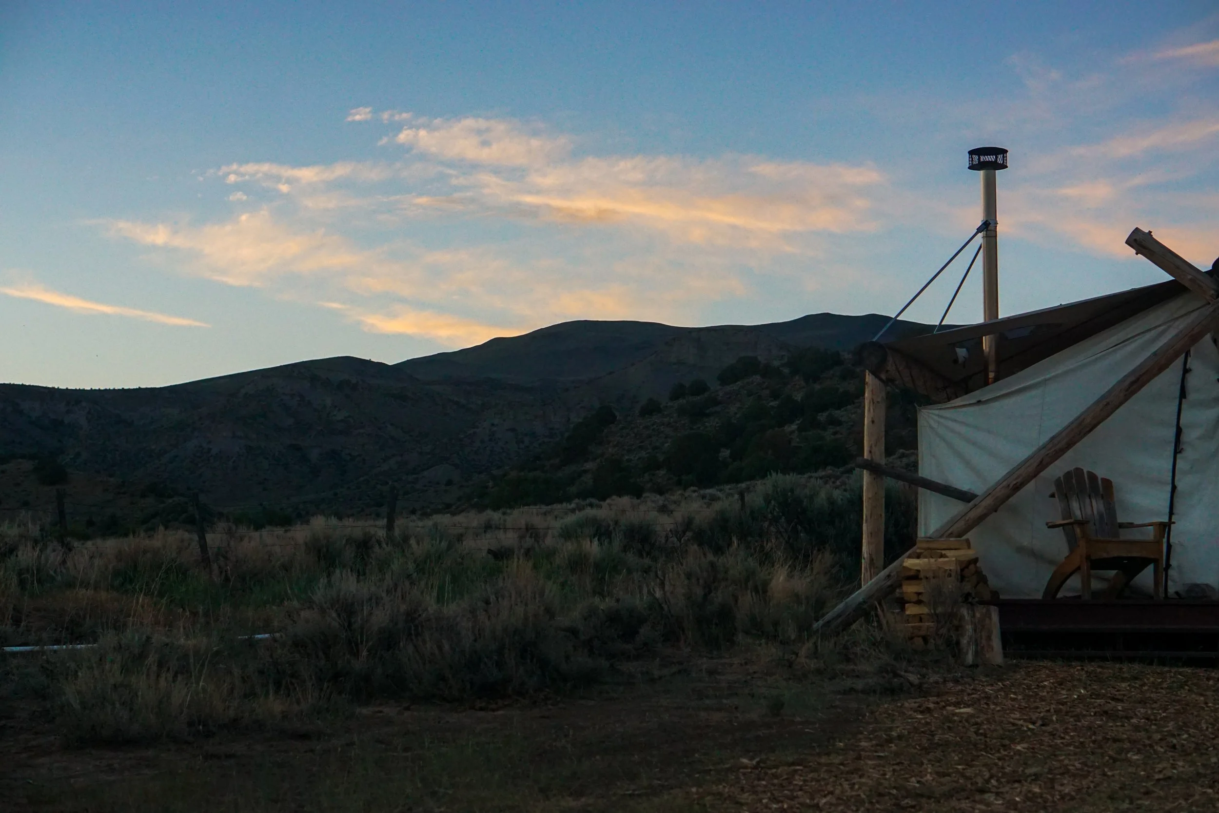A rustic outdoor canopy with a wooden chair on a porch, set against a mountain hillside during sunset with a partly cloudy sky.