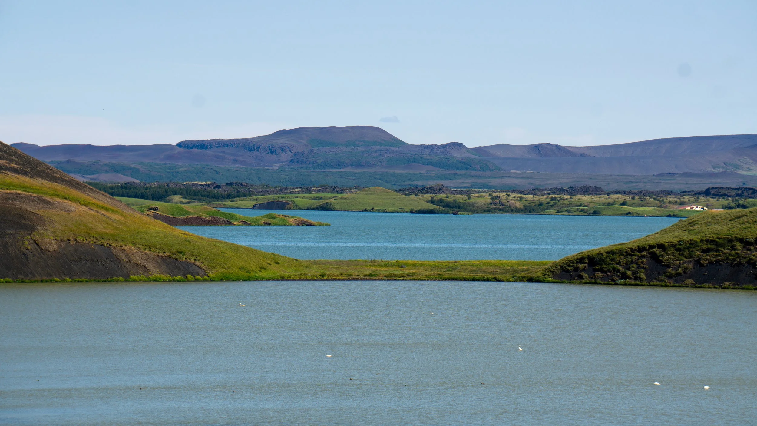 A scenic landscape with a lake in the foreground, surrounded by rolling green hills and mountains in the background, under a partly cloudy sky.