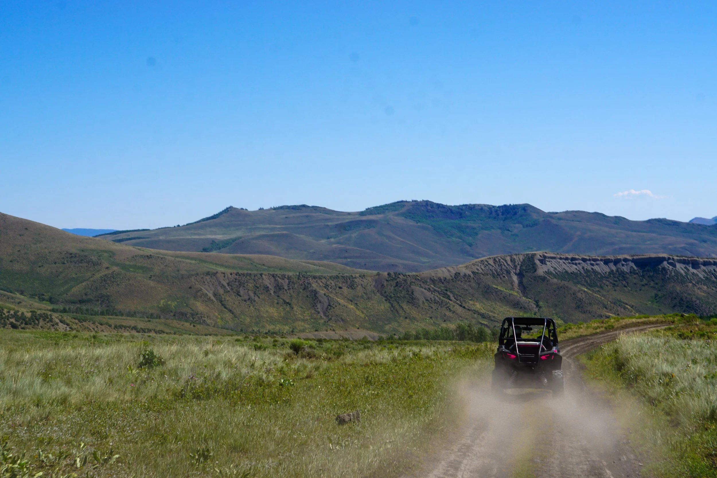 An off-road vehicle driving on a dusty dirt trail through a grassy plain with rolling hills and mountains in the background under a clear blue sky.