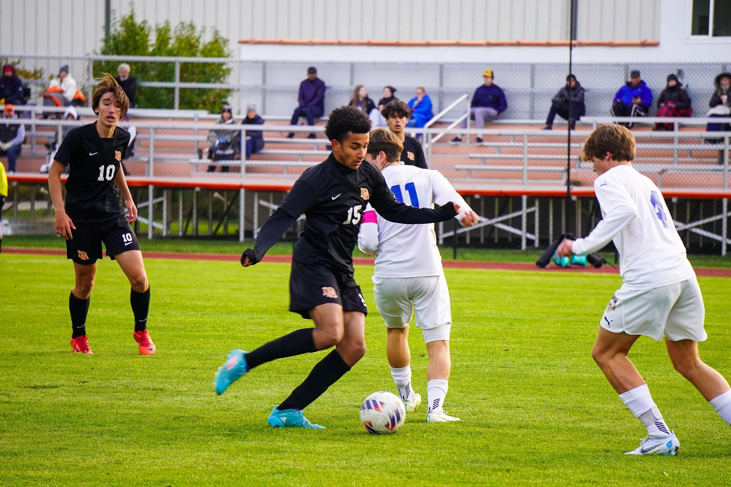 Soccer players competing for the ball on a grassy field, with spectators sitting in bleachers in the background.