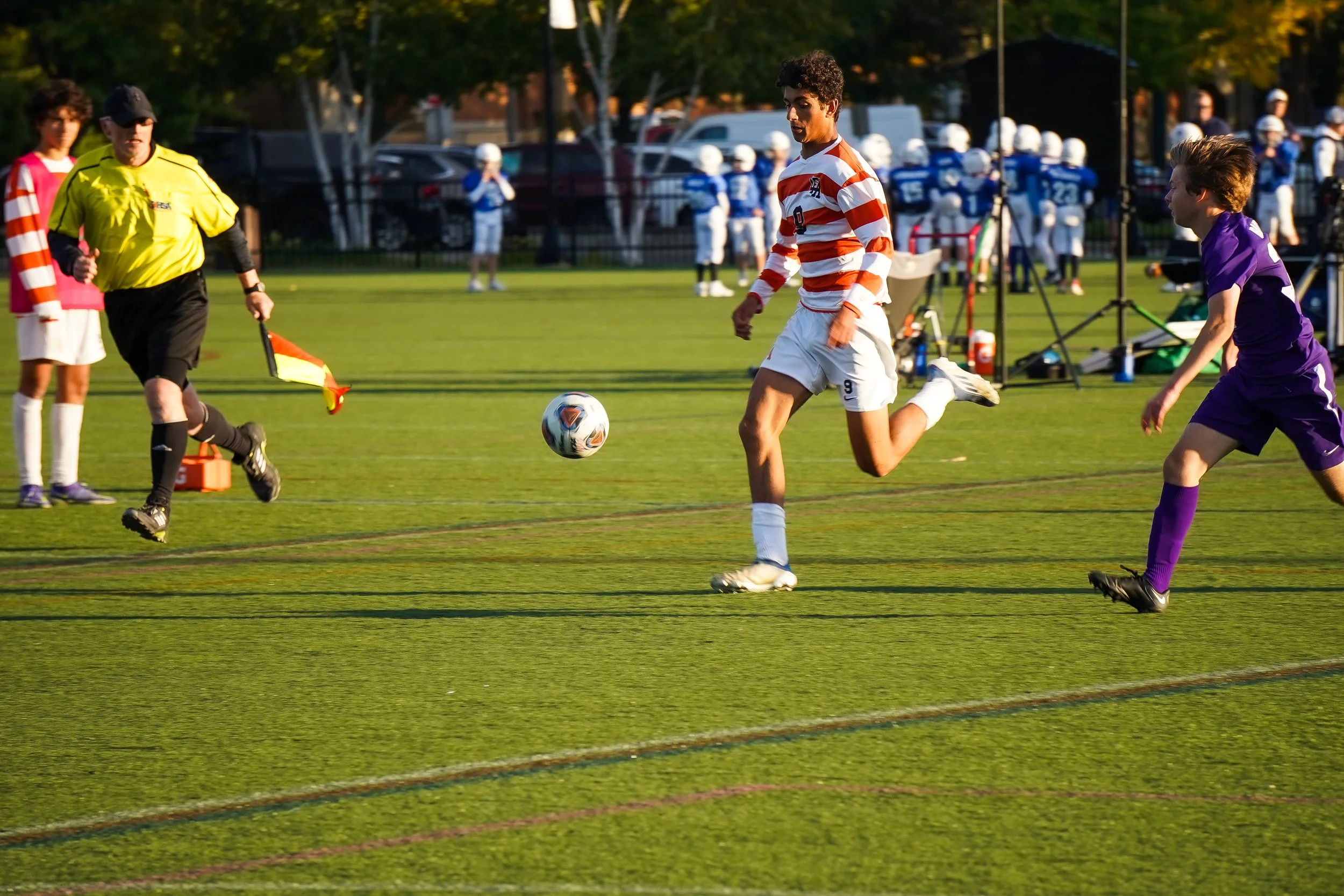 A soccer game with players, a referee, and a group of spectators in the background. A player in a red and white striped shirt is chasing the ball, while a player in purple is nearby. The referee is holding a yellow flag and running on the field.