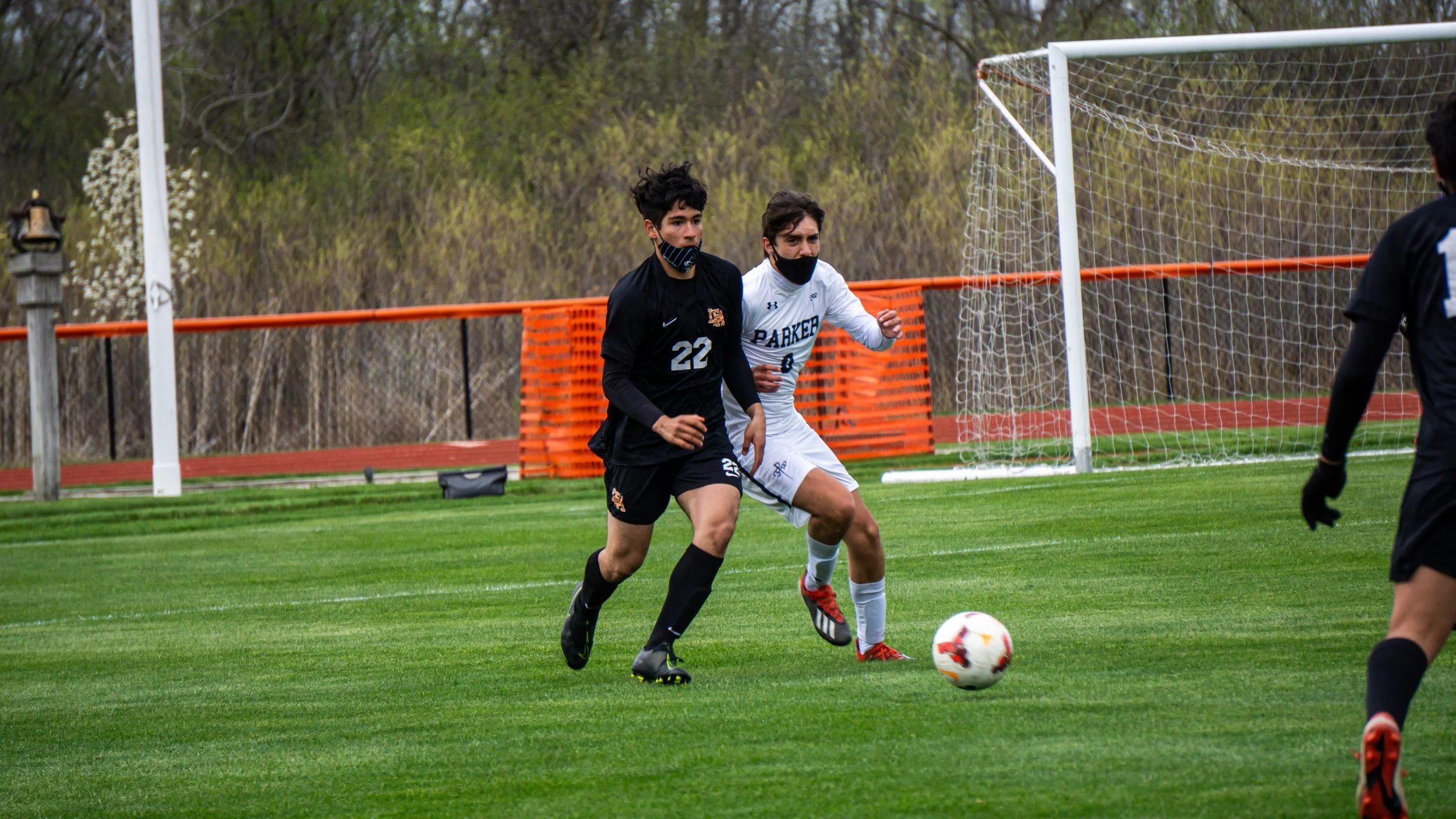 Two soccer players, one in black and the other in white, chase a soccer ball on a field. Both are wearing masks, and there is a goalpost behind them.