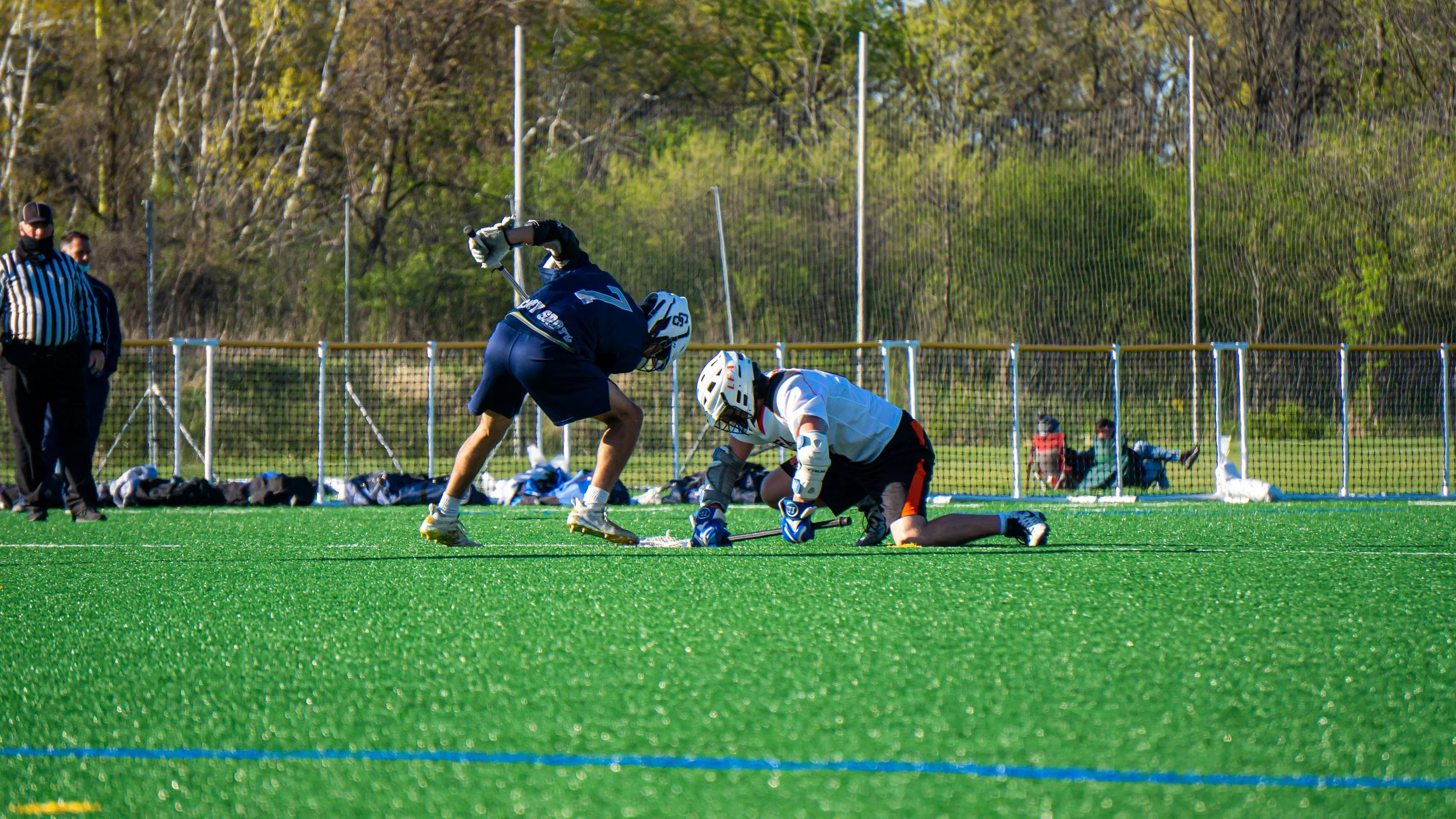Lacrosse game with players in helmets and pads, one kneeling on the turf and one standing, on a green field outdoors, with spectators behind a fence.
