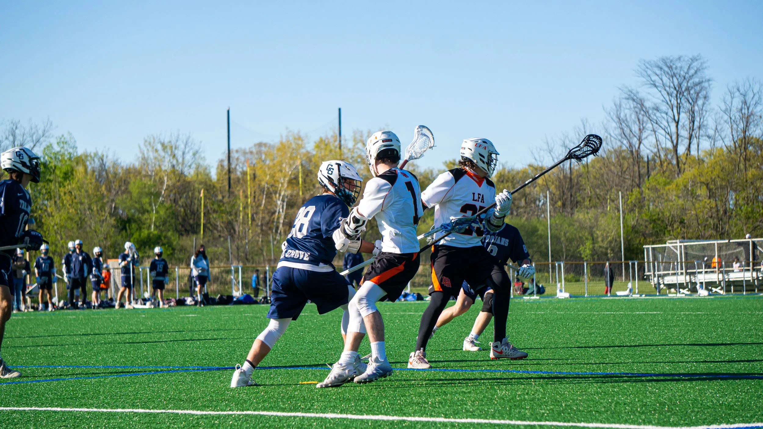 Lacrosse players competing on a green field under a clear blue sky, with players in white and navy uniforms and helmets, some holding lacrosse sticks, and a line of players and spectators in the background.