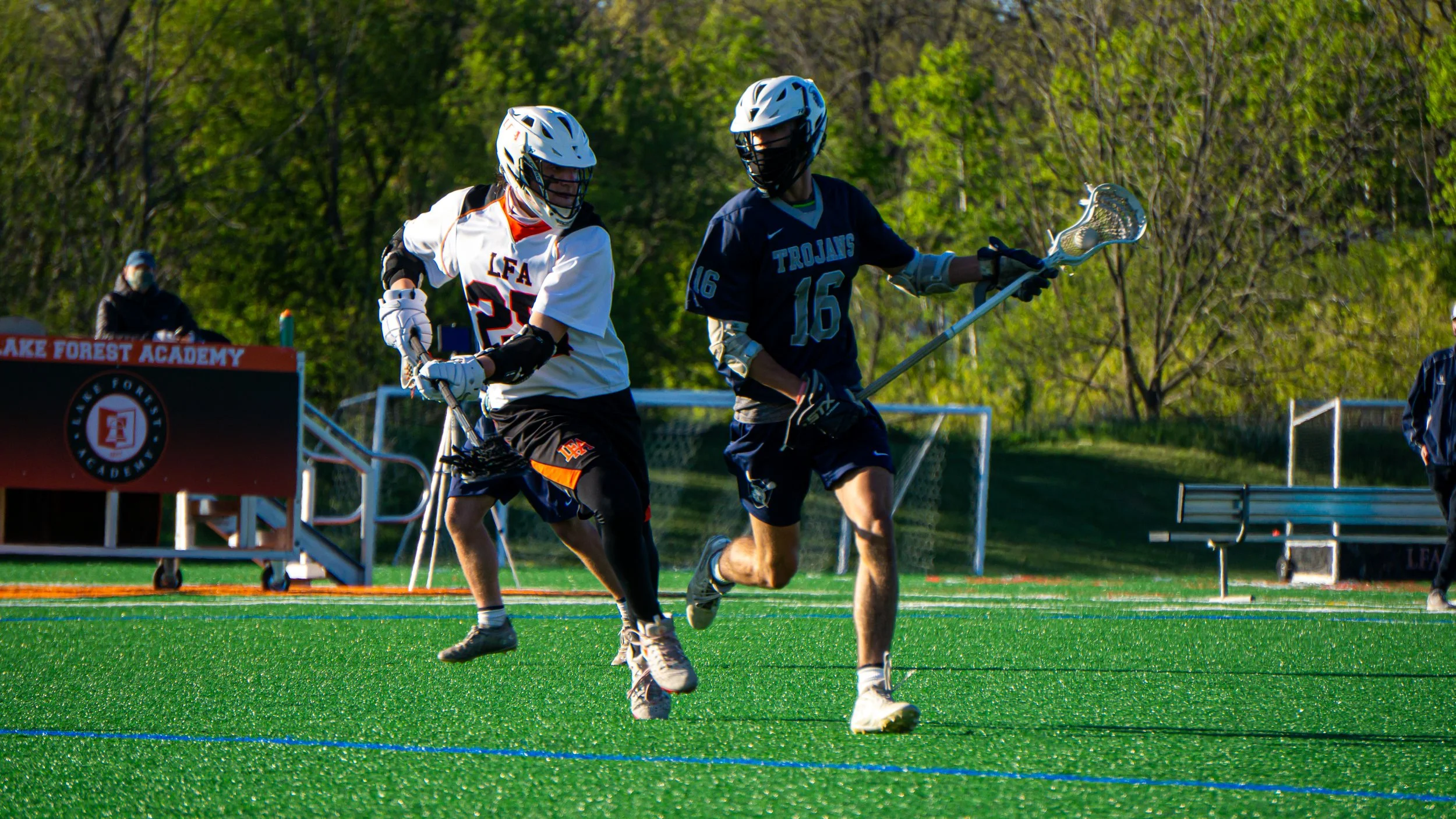 Two lacrosse players in action on a green field, with one player wearing a white jersey and the other in a blue jersey, both wearing helmets and protective gear, holding lacrosse sticks, with trees and a sign in the background.