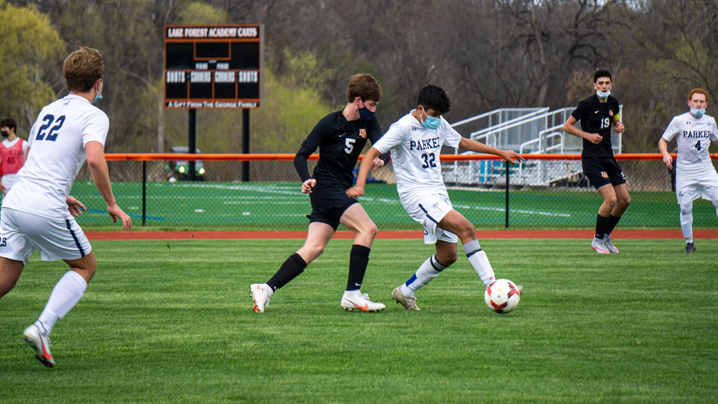 Soccer game with players in black and white uniforms, all wearing face masks, on a green field during daytime.