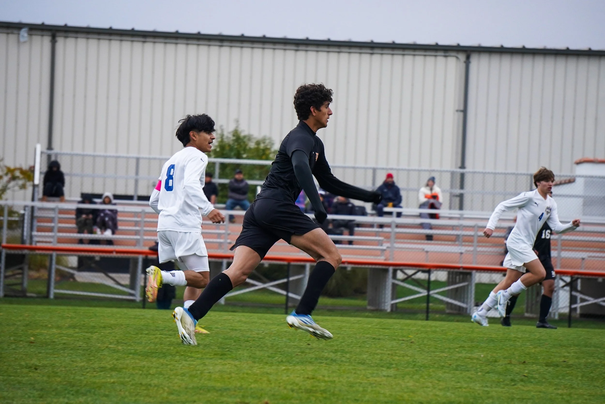 Soccer players running on the field during a game, with spectators sitting on bleachers in the background.