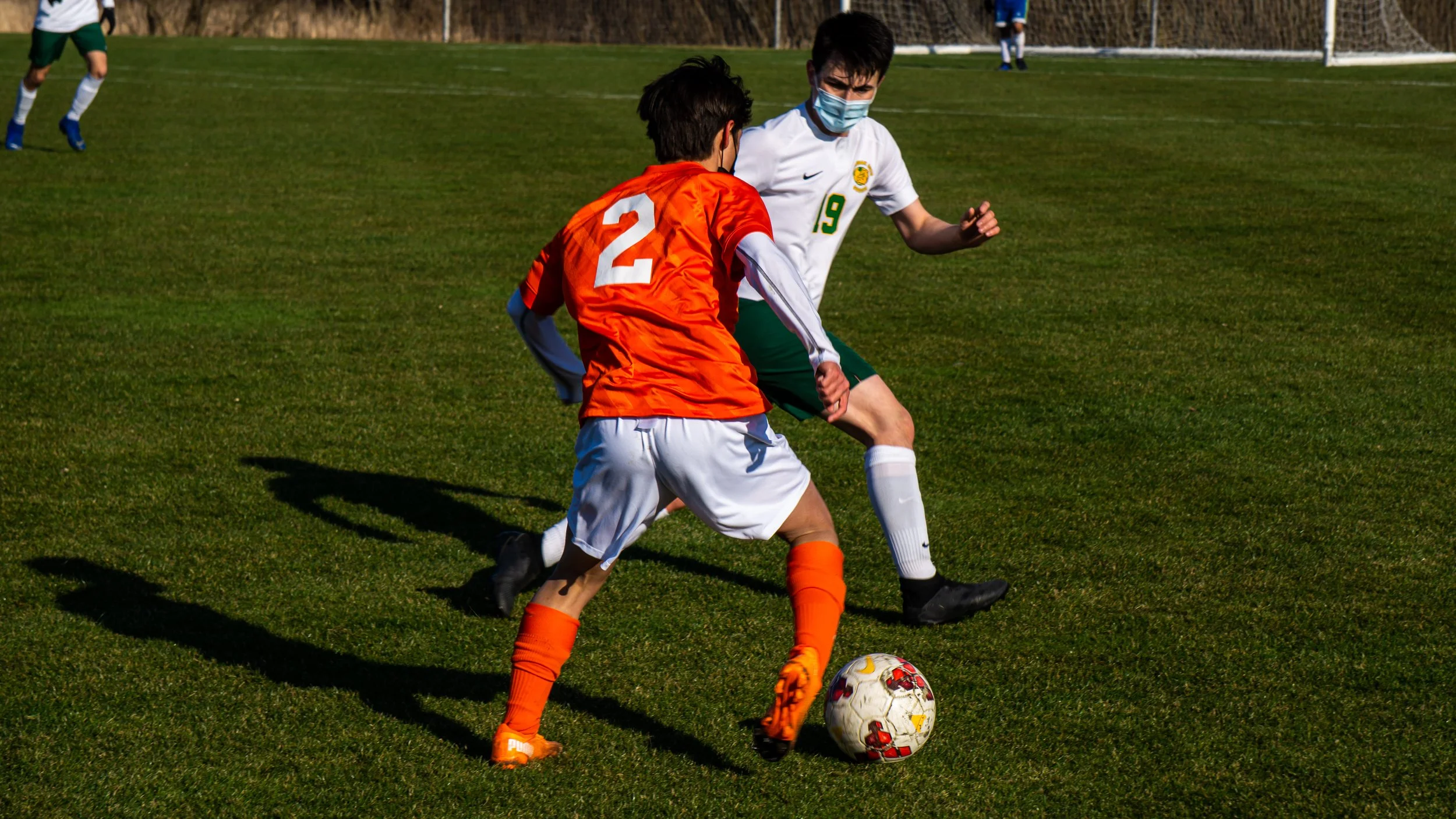 Two young soccer players, one in an orange jersey and the other in a white jersey, compete for the ball on a green field during daytime. Both players are wearing face masks.