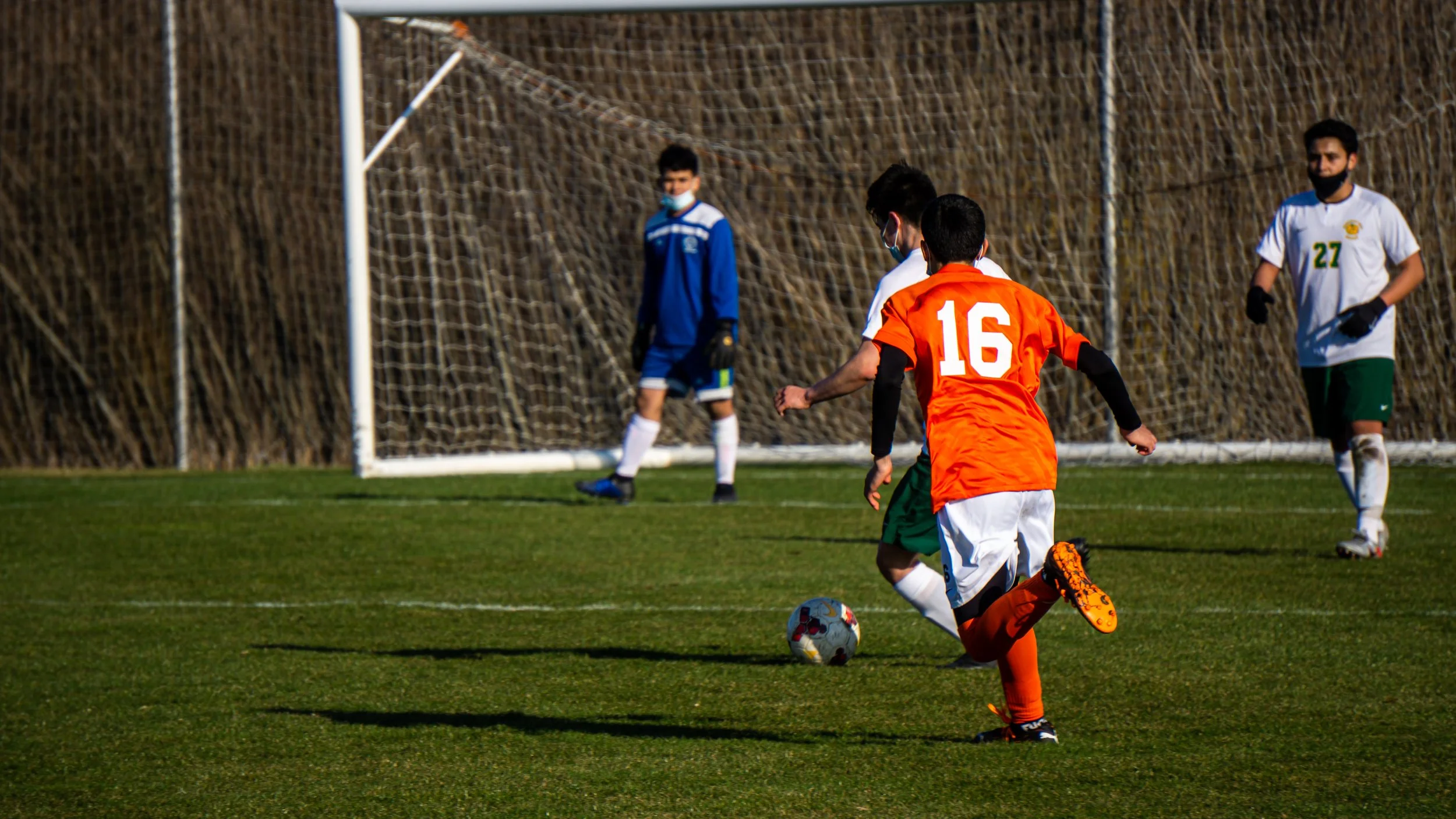 Soccer match with a player in orange jersey number 16 kicking the ball near the goal, while two players in white and one in blue are nearby on the field.