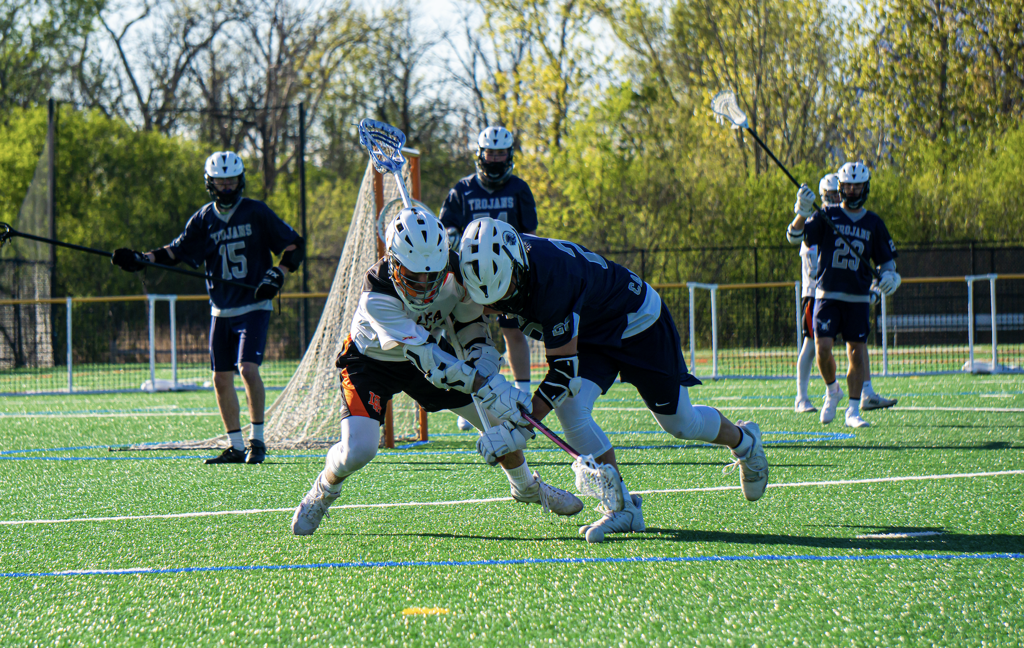 Lacrosse players in blue uniforms and helmets competing for the ball on a green field, with one player in white trying to gain possession, and others in the background holding lacrosse sticks.