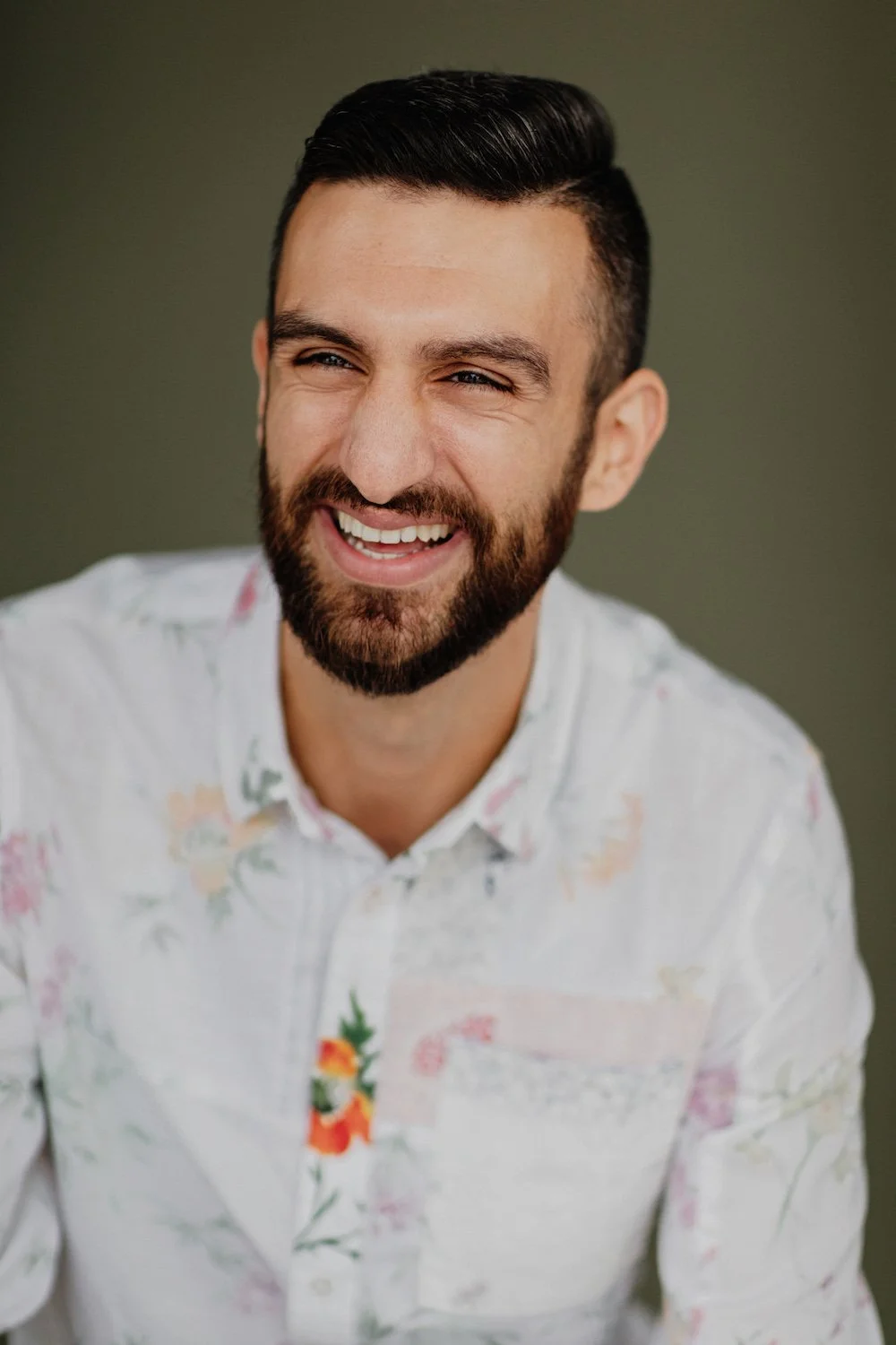 A smiling man with dark hair and a beard, wearing a white floral shirt, against a dark green background.