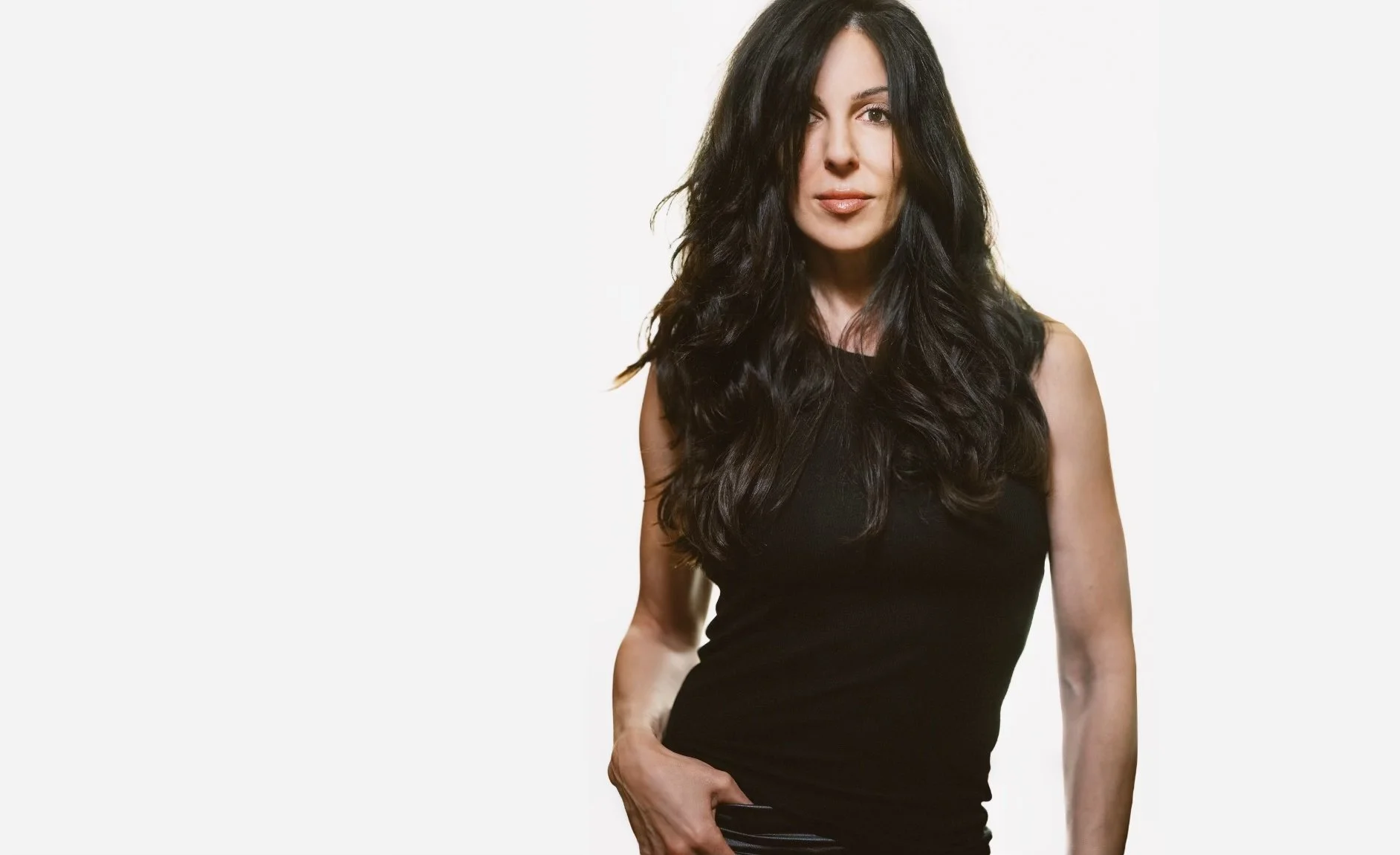 A woman with long black hair wearing a sleeveless black top standing against a plain white background.