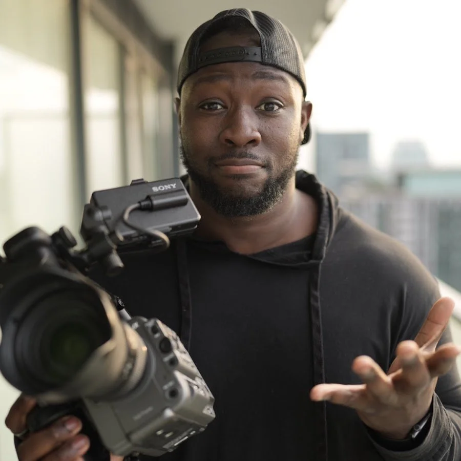A man wearing a black hoodie and a backward black cap, holding a professional camera, standing on a balcony with city buildings in the background, gesturing with his right hand.