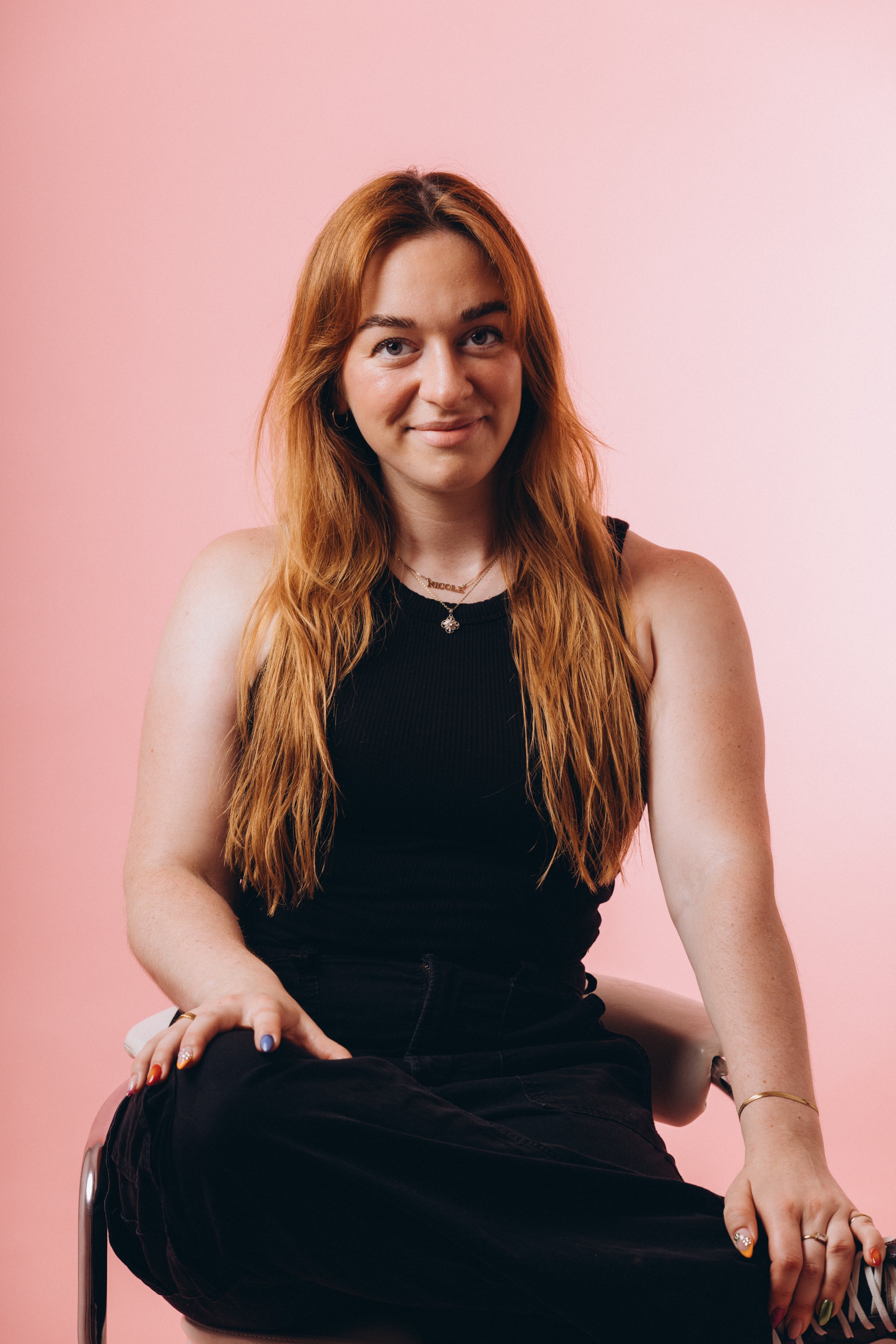 A young woman with long red hair, wearing a black sleeveless top, sitting against a pastel pink background.