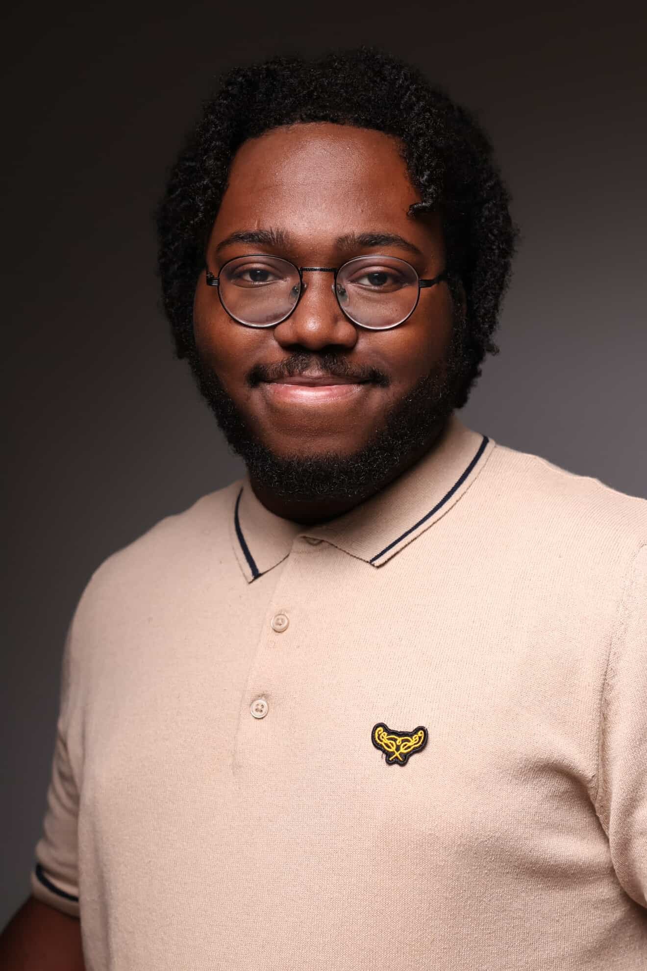 Portrait of a young man with glasses, a beard, and curly hair, wearing a beige polo shirt with a black embroidered logo, smiling against a dark background.