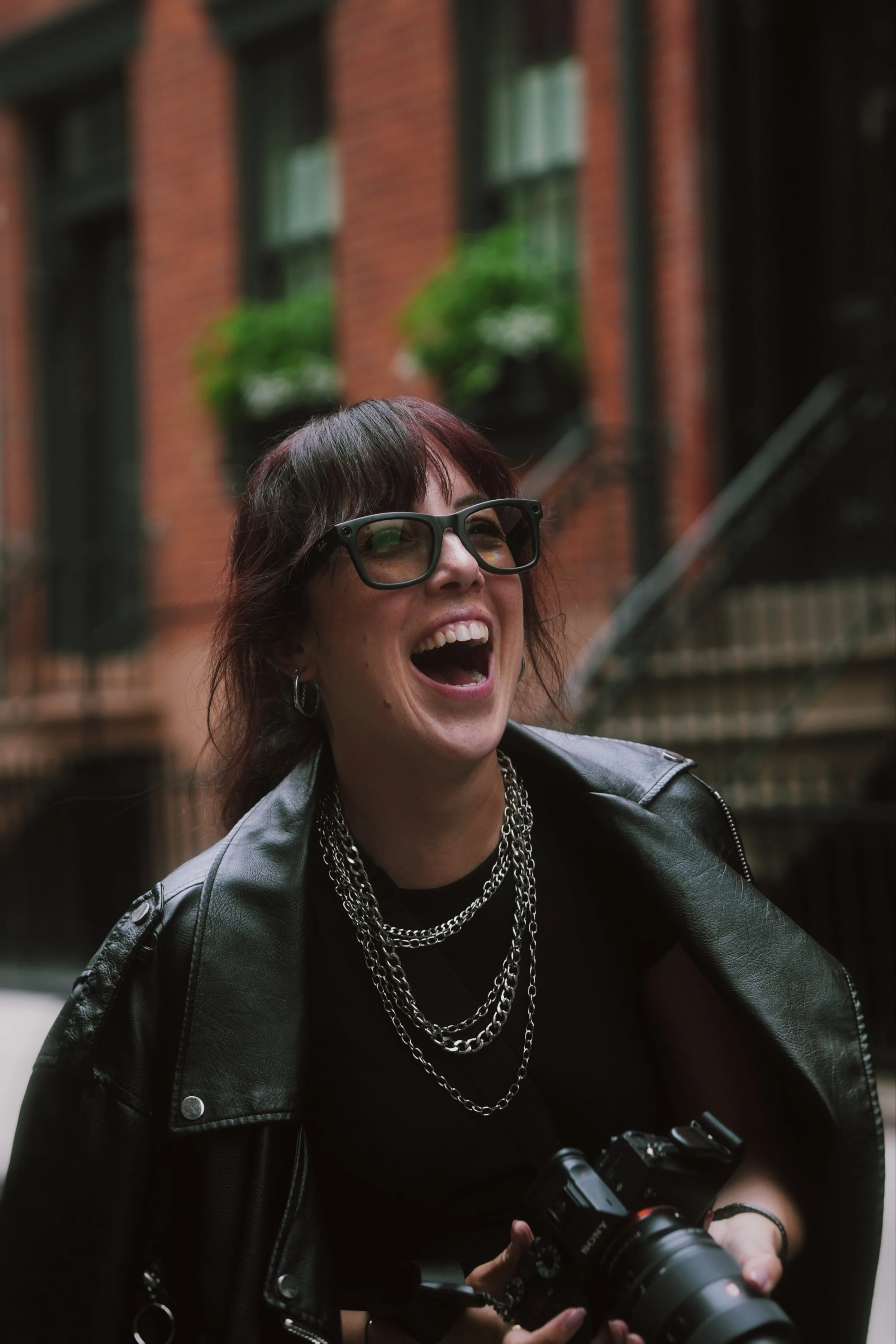 Woman with short dark hair, glasses, leather jacket, and layered chains, smiling while holding a camera on a city street.