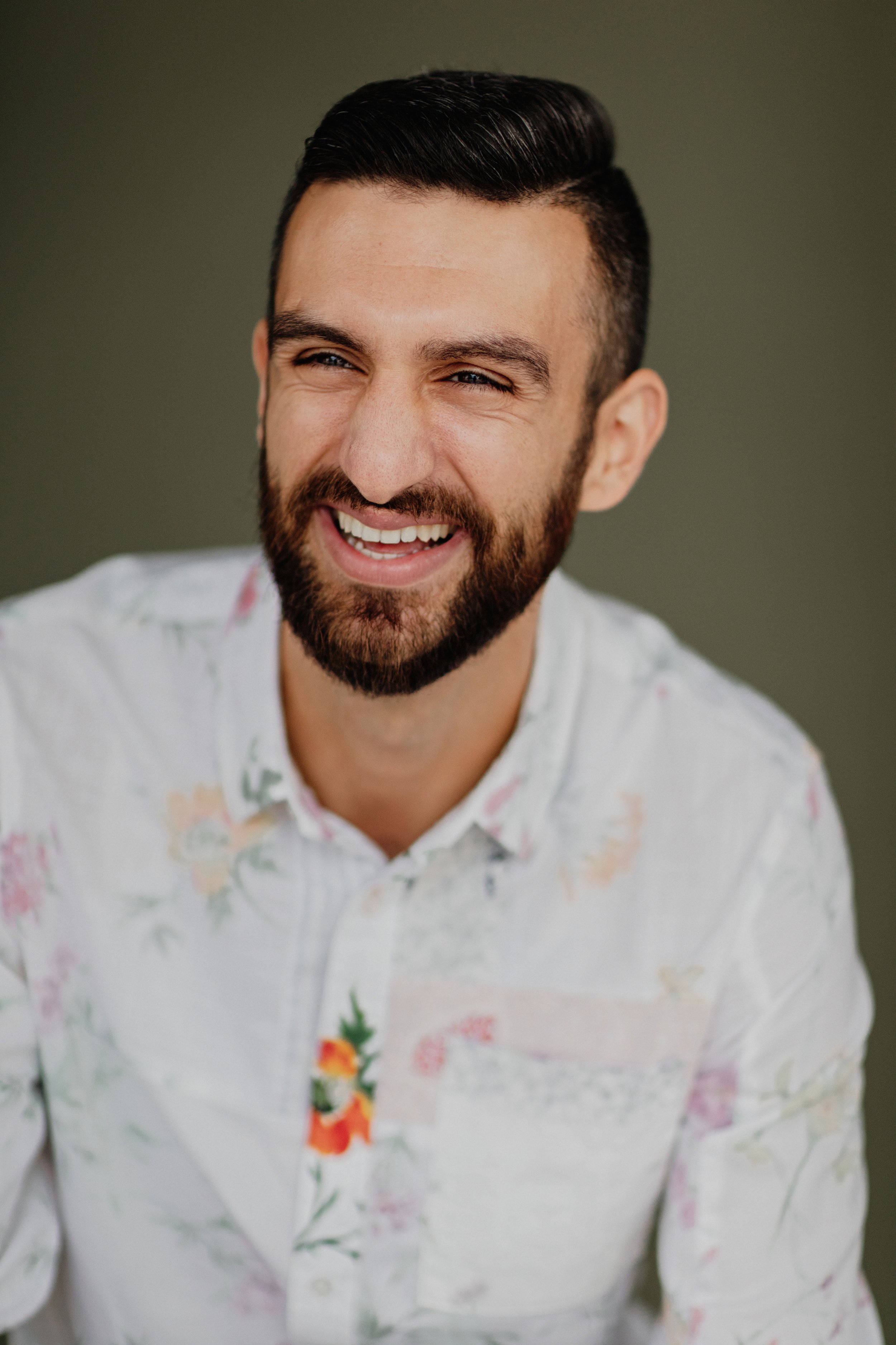 A man with dark hair and a beard smiling and looking to the side, wearing a white floral shirt, against a dark green background.