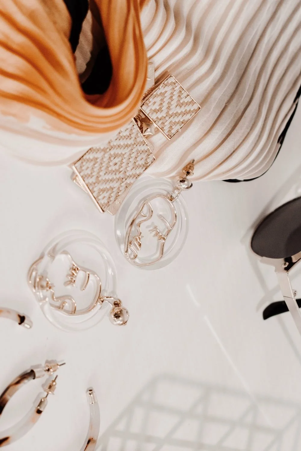 Close-up of fashionable accessories including woven gold earrings, transparent earrings with face designs, and a striped fabric background.