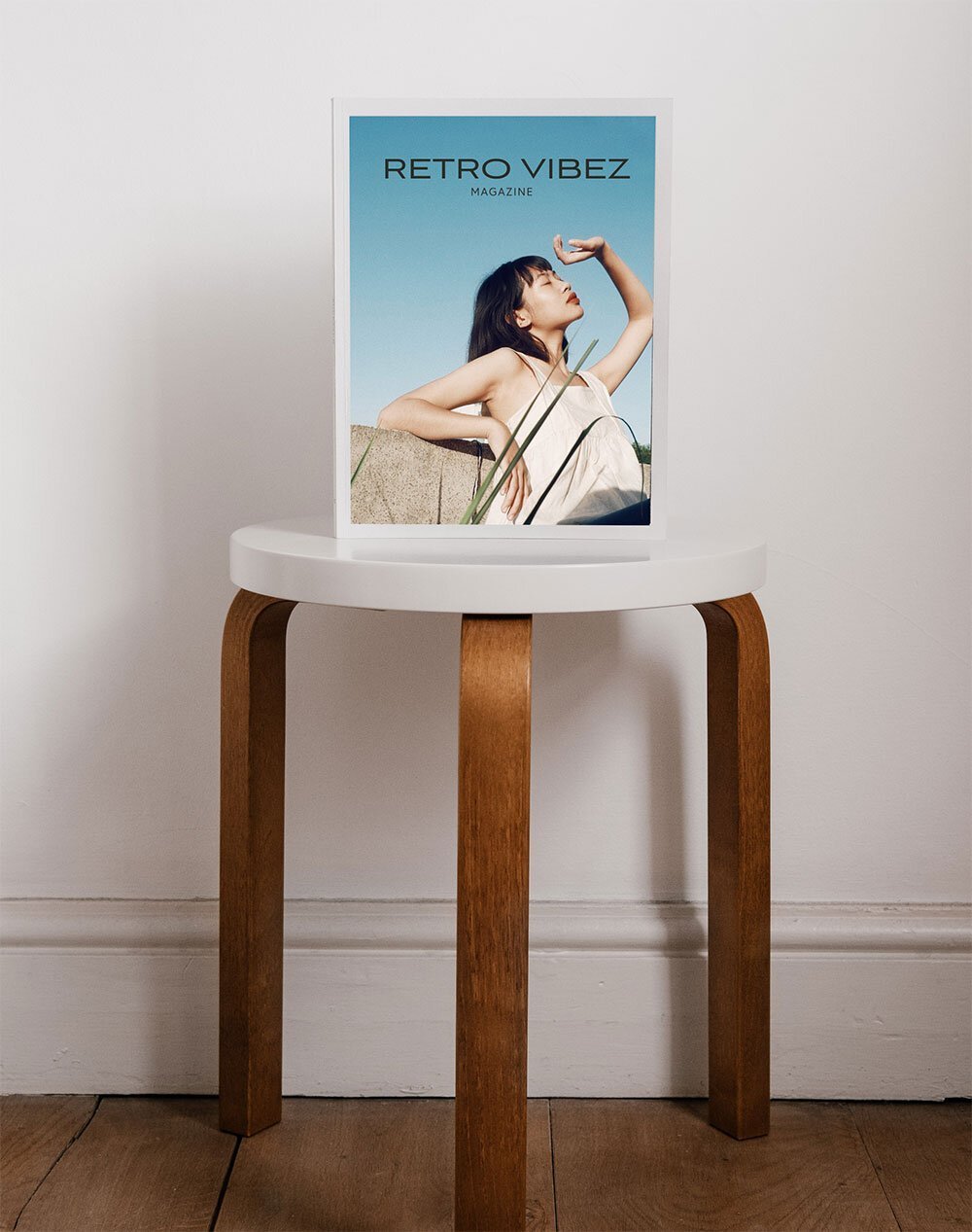 Magazine titled "Retro Vibez" on a round white table with wooden legs, featuring a woman in a white dress with black hair, posing outdoors against a blue sky background.