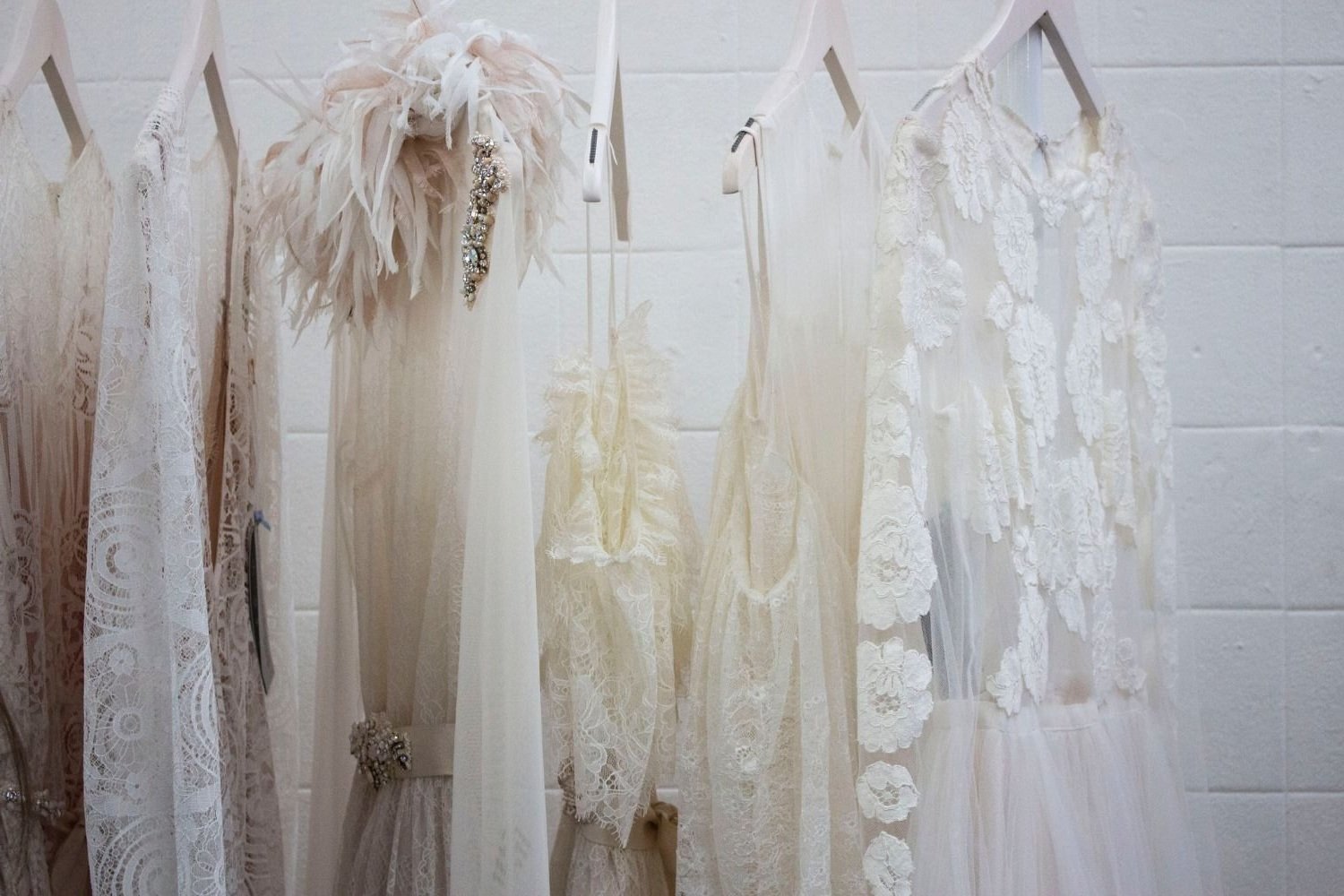 A row of cream and white lace and fabric dresses hanging on a rack against a white tiled wall, with some dresses featuring feather, floral lace, and embellishments.