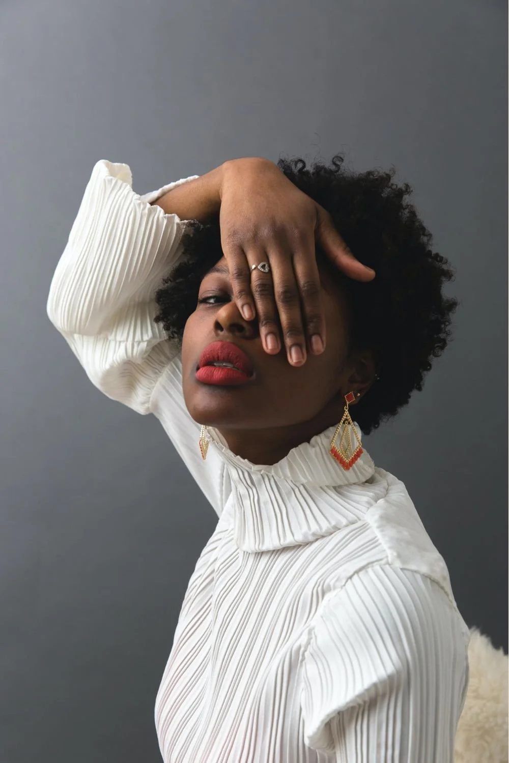 A woman with curly black hair wearing a white pleated shirt, red lipstick, and colorful earrings, covering her eye with her hand against a gray background.