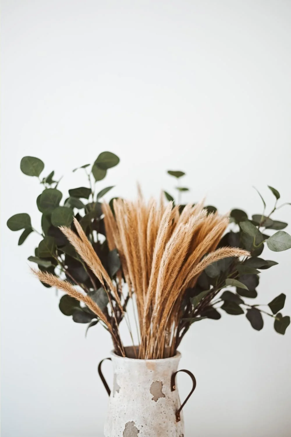 A white ceramic vase with metal handles holding a bouquet of green eucalyptus leaves and beige pampas grass.