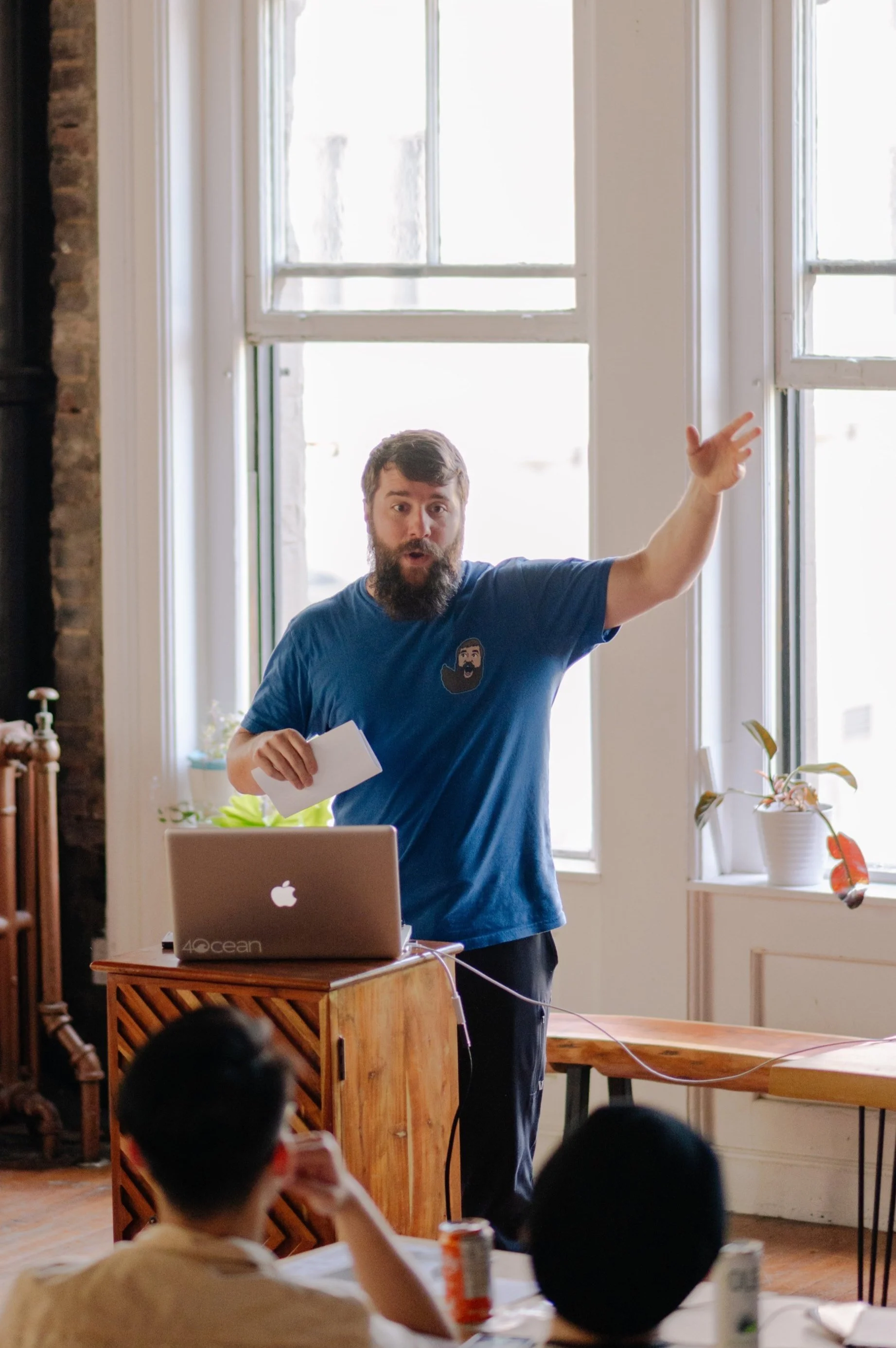 A man with a beard and short hair, wearing a blue t-shirt, is giving a presentation in a bright room with large windows. He is gesturing with his right hand while holding a piece of paper in his left hand. There is an open laptop on a wooden stand in front of him and a group of people watching.