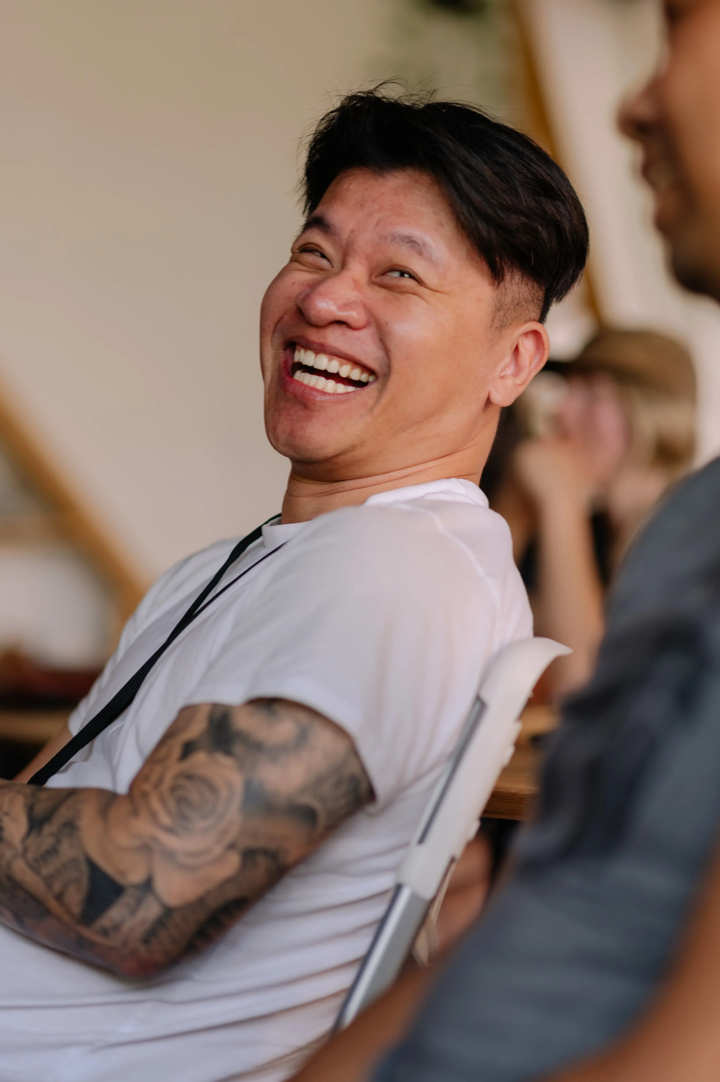 A man with short black hair, tattoos on his arm, and a white t-shirt, smiling and laughing while sitting in a chair, with other people blurred in the background.