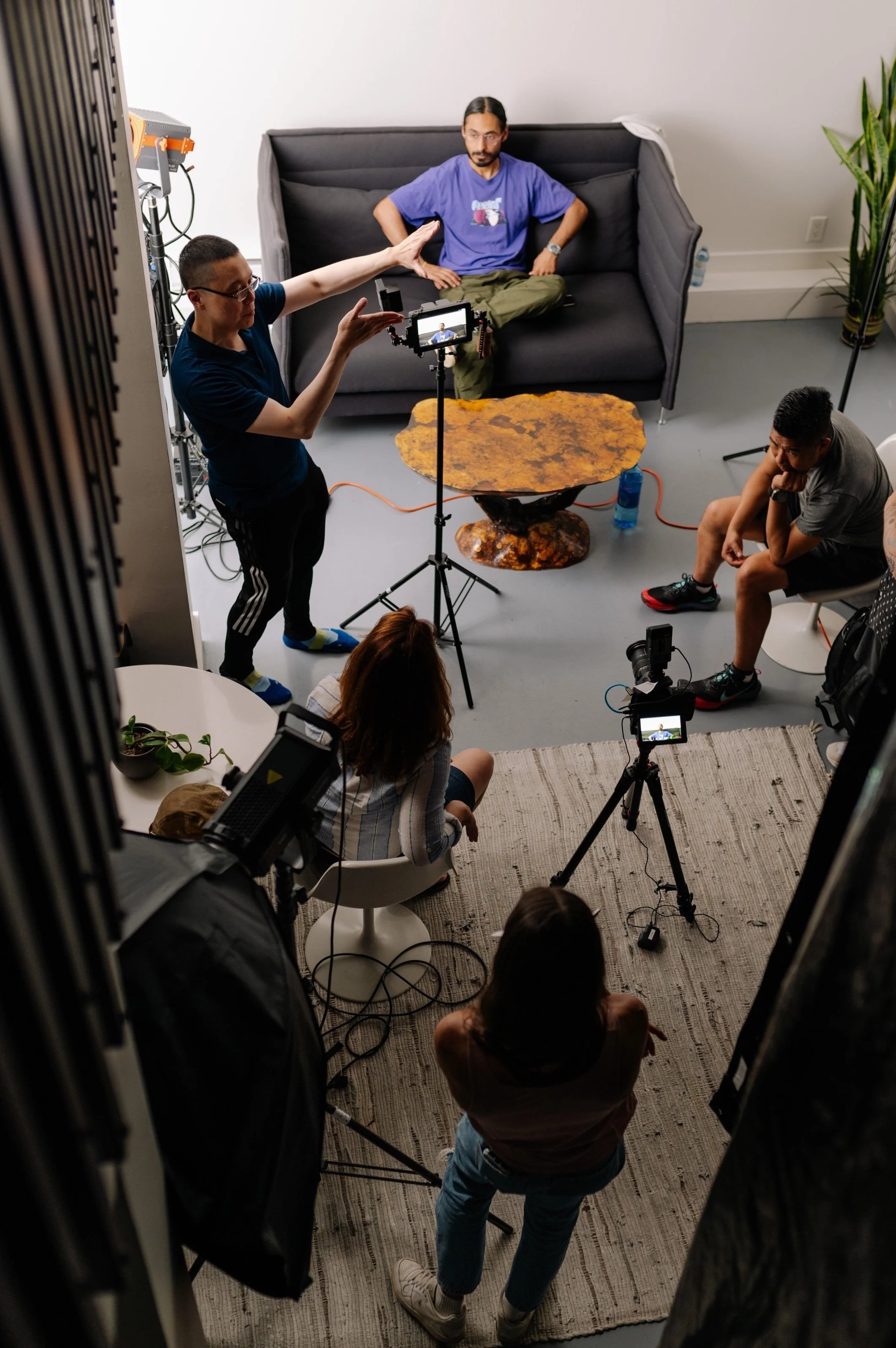 A behind-the-scenes photo of a video or photo shoot in a room with a gray couch, a rustic wooden coffee table, and several people including a woman sitting, a man sitting on the floor, a woman standing, and a man standing near camera equipment.