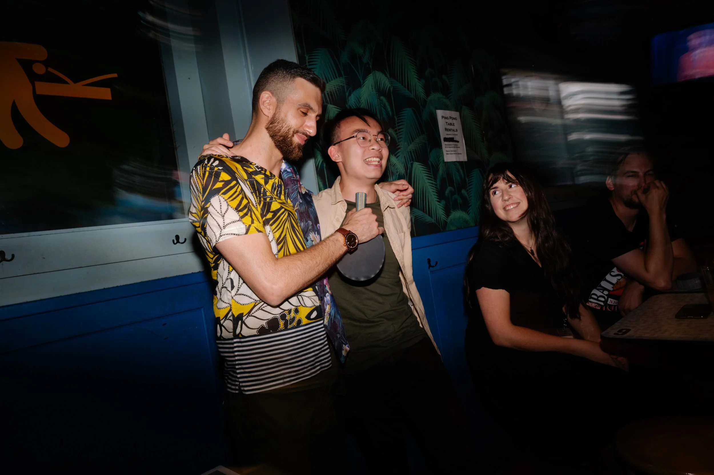 Four young adults sitting at a bar or club, with two men standing close together, one holding a ping pong paddle, and two women sitting and smiling. The background features dark tropical-themed decor.