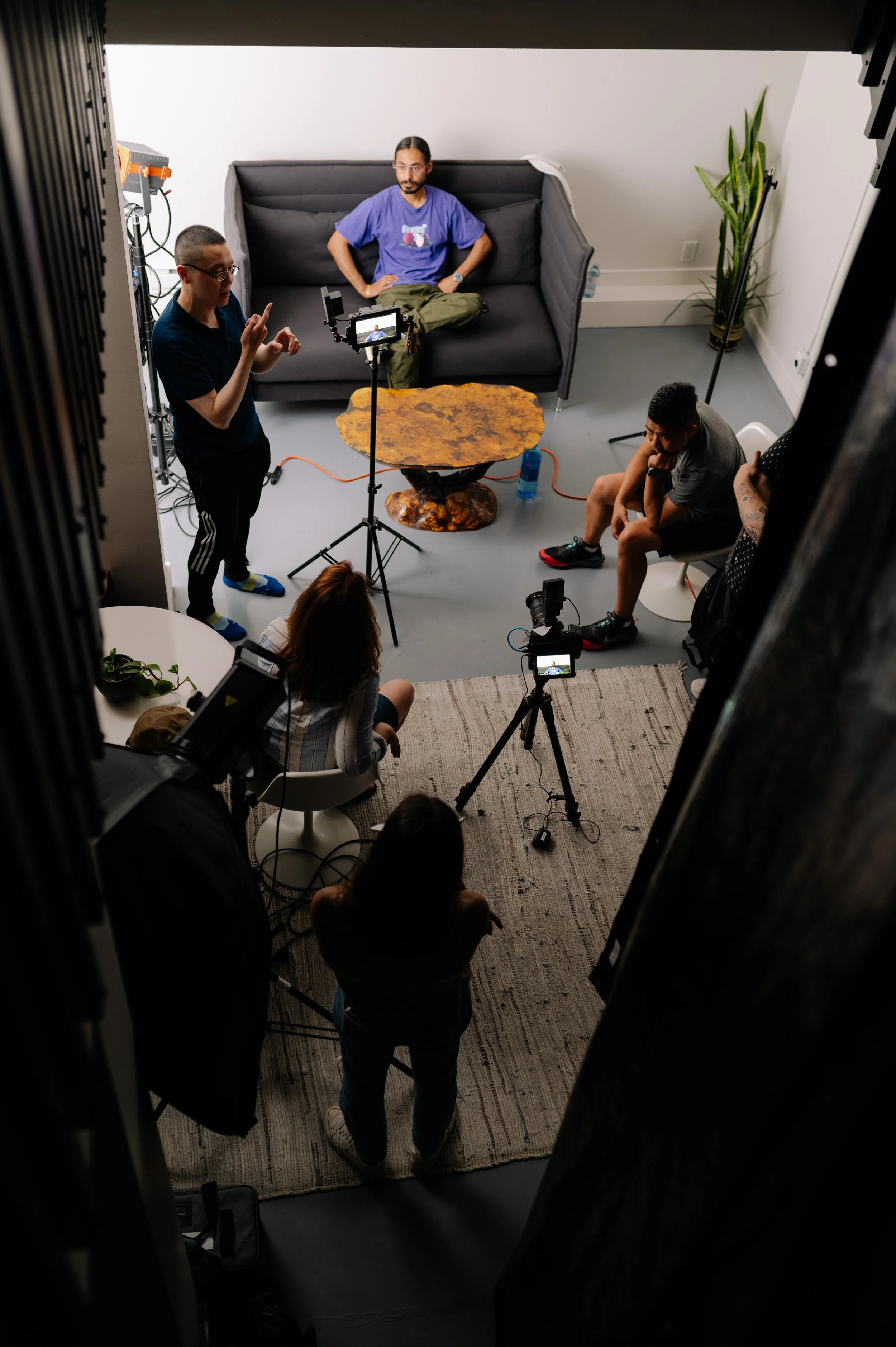 A small group of people are filming an interview scene in a cozy indoor setting with a dark sofa, a round marble table, and a plant in the corner. There are two cameras and a lighting setup, with three individuals positioned around the scene, one person standing and speaking, and the others seated or focused on recording.