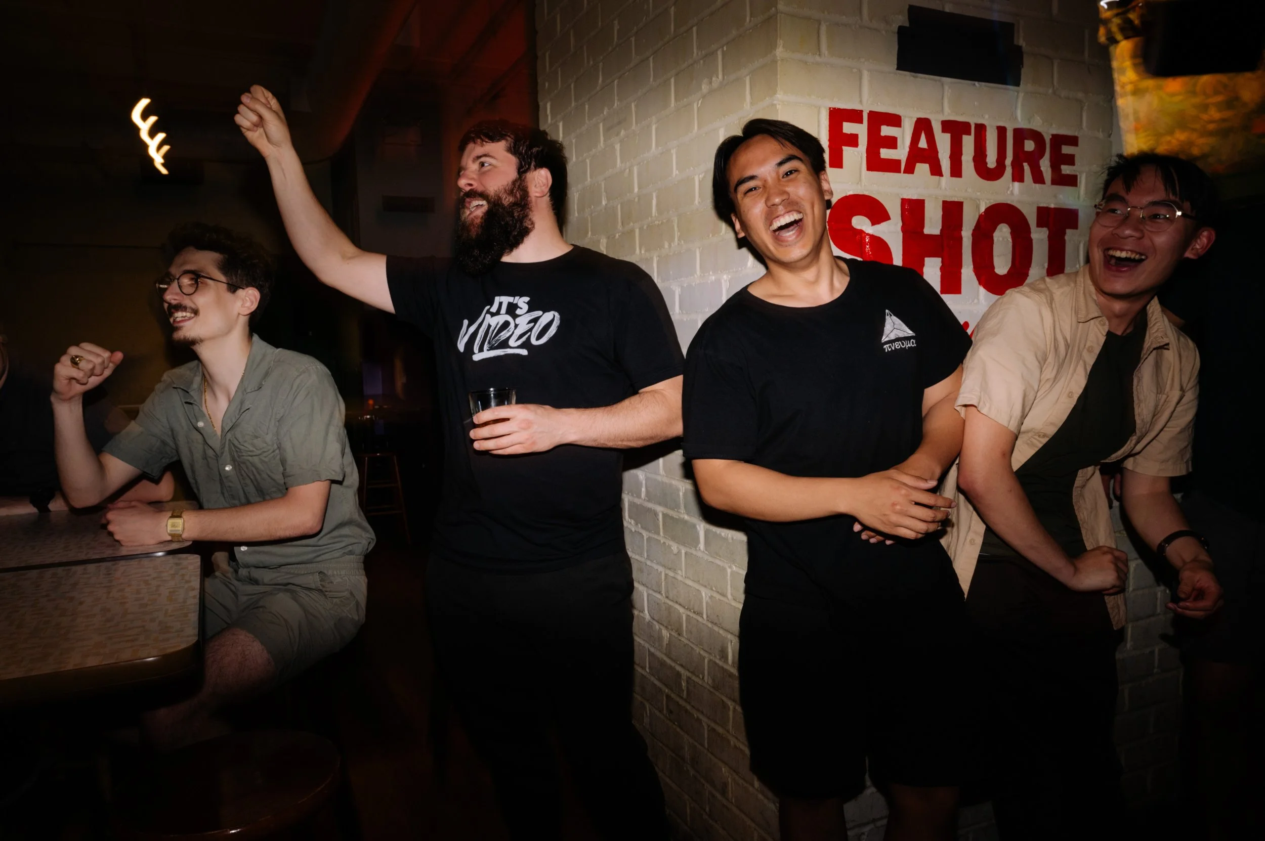 A group of people celebrating and laughing at a bar or nightclub, with a white brick wall and a red sign reading 'FEATURE SHOT' in the background.