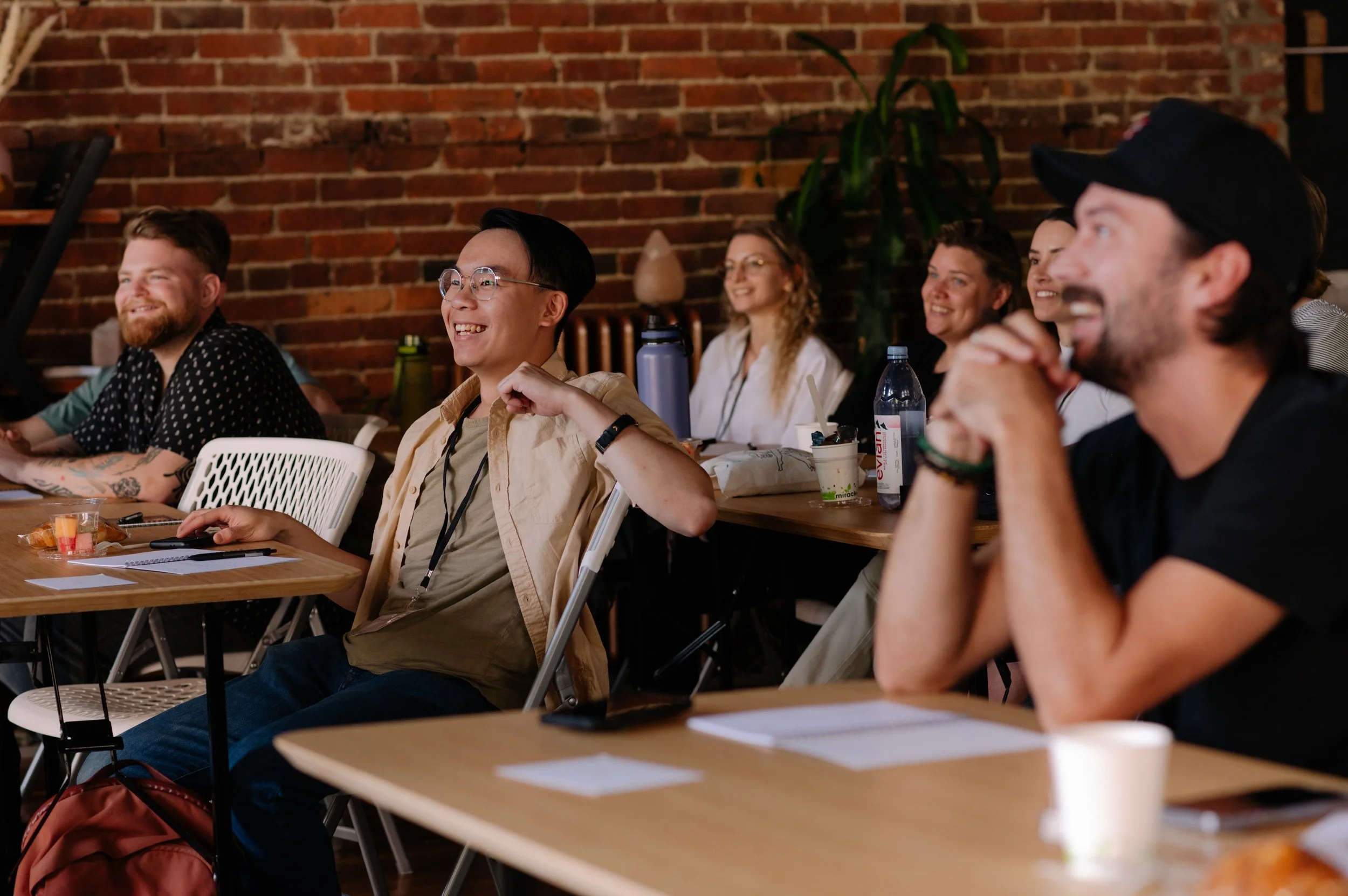 Group of diverse young adults sitting at tables in a brick-walled room, attending a seminar or workshop, smiling and engaged.