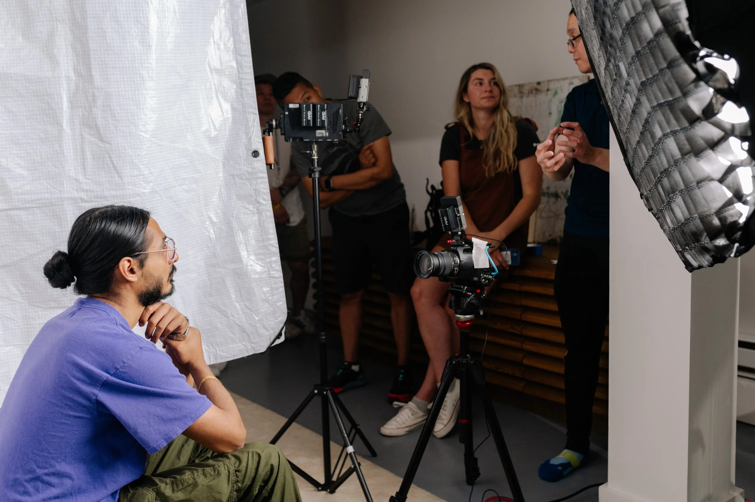 A man with glasses and a man bun wearing a purple shirt is sitting on the floor, looking at a scene being filmed in a studio. There are several people behind, including women with long hair, some with cameras, and a woman holding a makeup sponge, pre